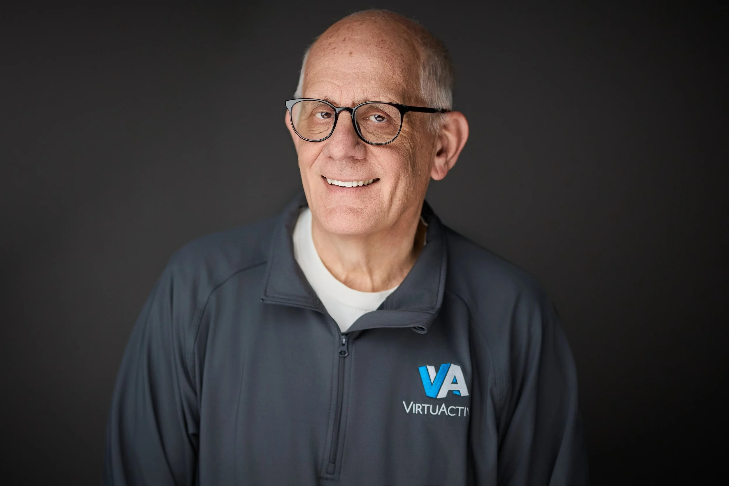 A smiling man with short, gray hair and a beard, wearing a black shirt, standing in front of a colorful geometric background.
