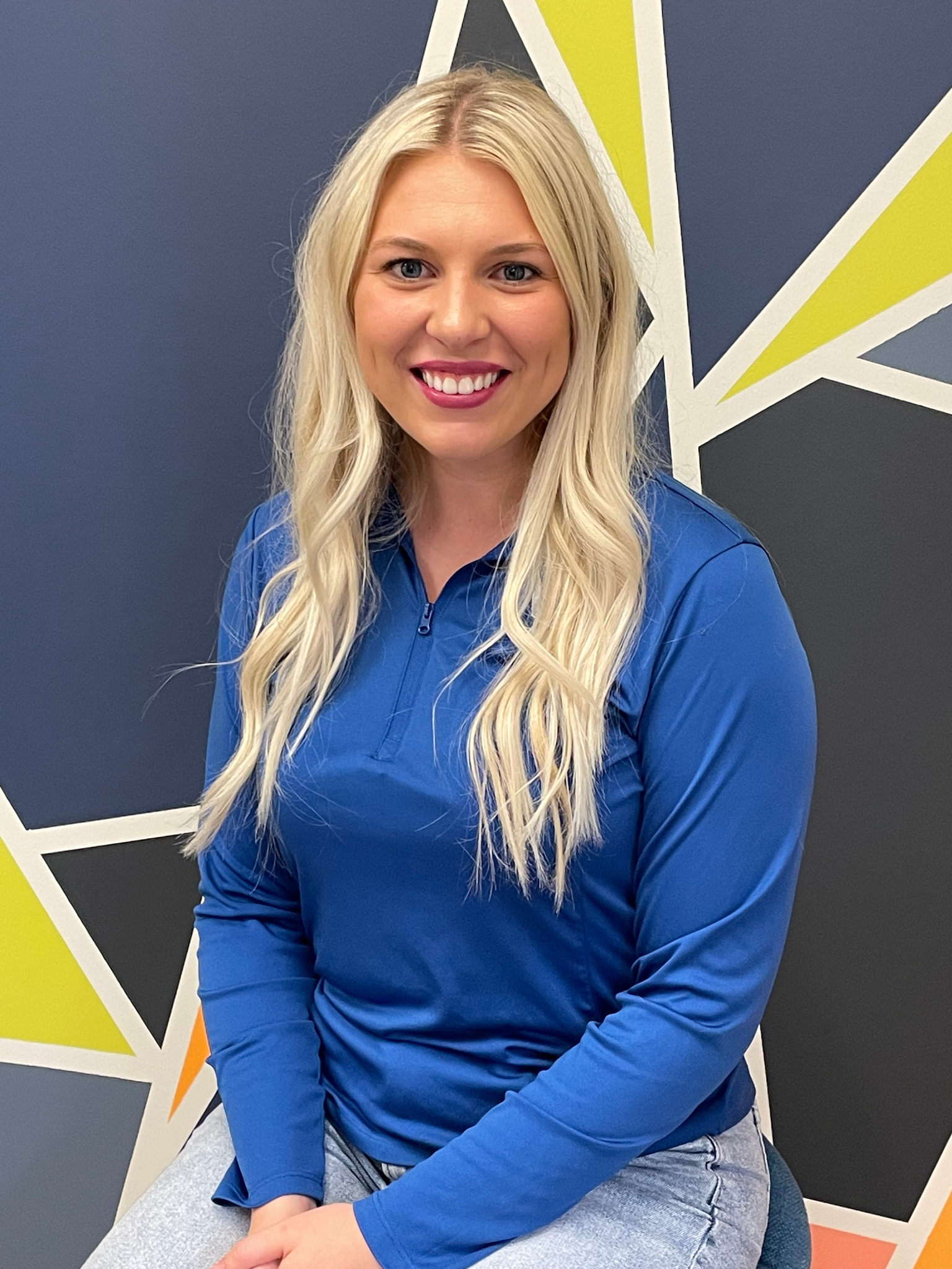 A woman with long blonde hair wearing a blue zip-up shirt, smiling at the camera, standing in front of a colorful geometric wall design.