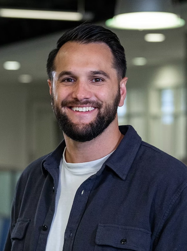 A smiling man with a beard and dark hair, wearing a dark shirt over a white T-shirt, standing in an office environment with blurred background lighting and windows.