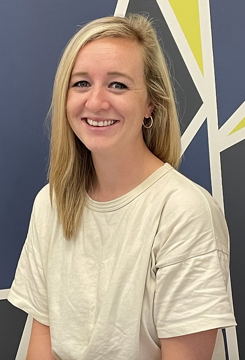A woman with long blonde hair smiling, wearing a beige T-shirt, standing in front of a geometric painted wall.