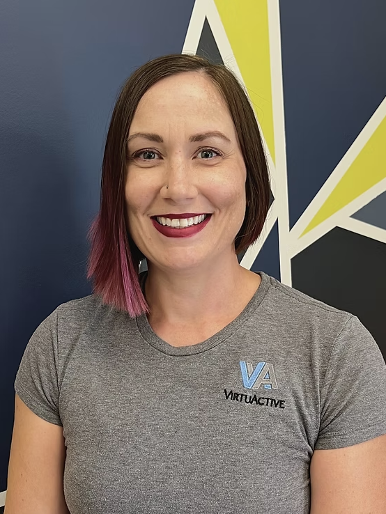 A smiling woman with short brown hair and purple-tinted tips, wearing a gray 'Virtuactive' t-shirt, standing in front of a wall with geometric designs in blue, yellow, white, and black.
