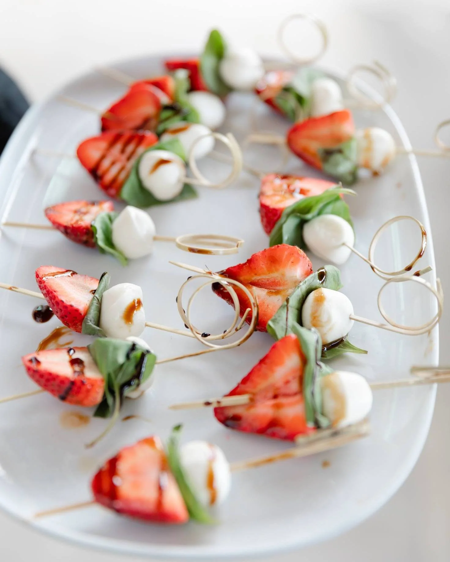 Skewers with strawberries, mozzarella balls, basil leaves, and drizzle of balsamic glaze on a white platter.