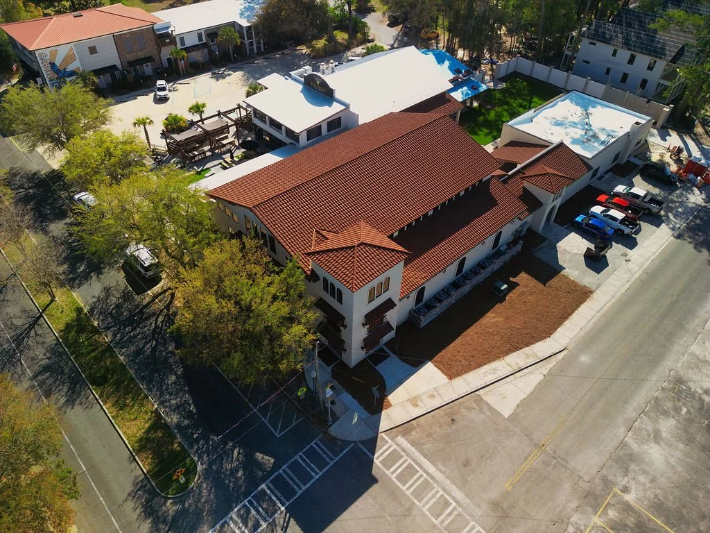 Aerial view of a building with a red tile roof, surrounded by parking lot, trees, and neighboring structures.