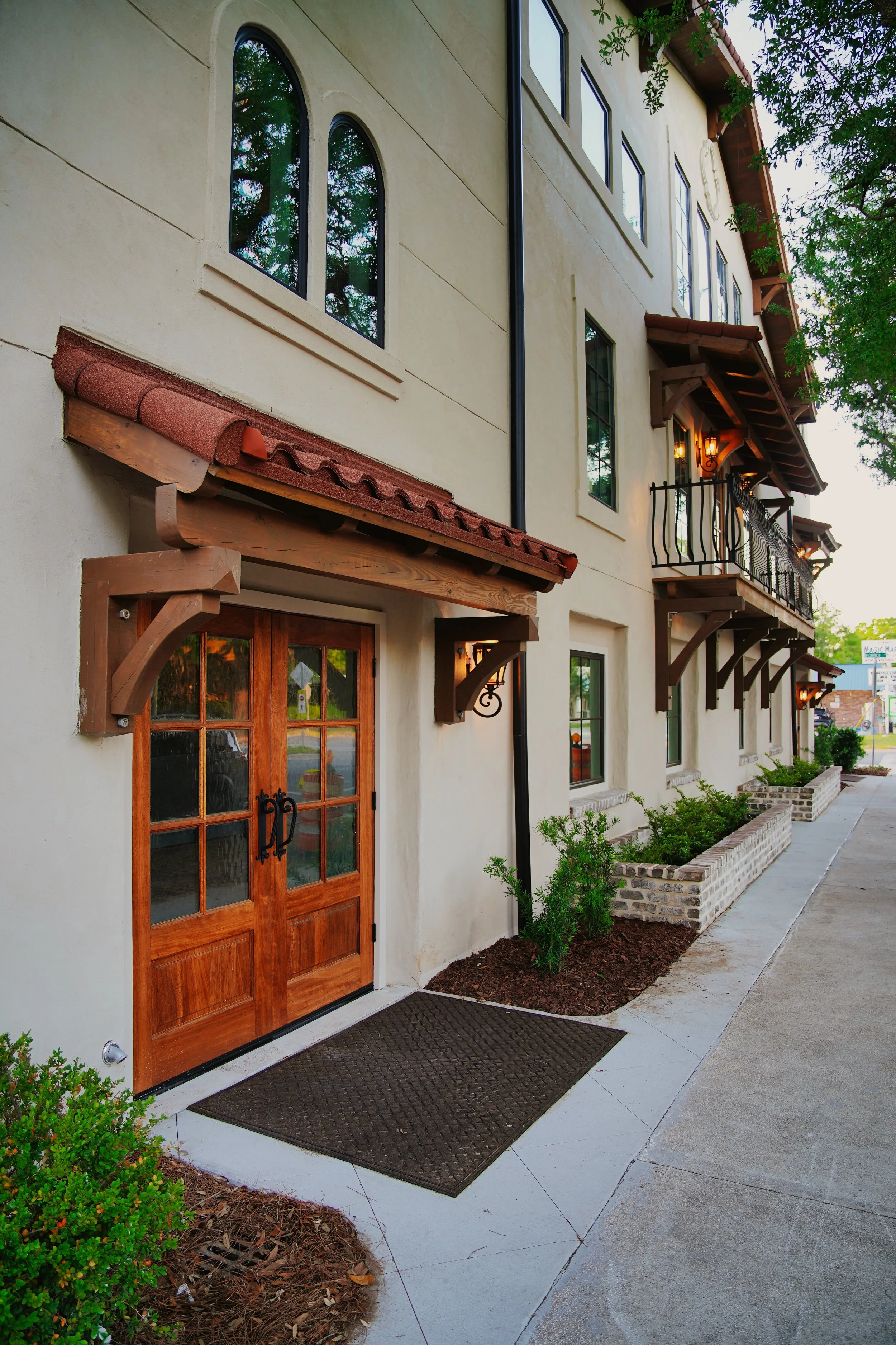 Exterior view of a multi-story building with beige stucco walls, red-tiled awnings above wooden doors, and black iron balconies on the upper floors. There are small landscaped garden beds with shrubs and a concrete sidewalk in front.