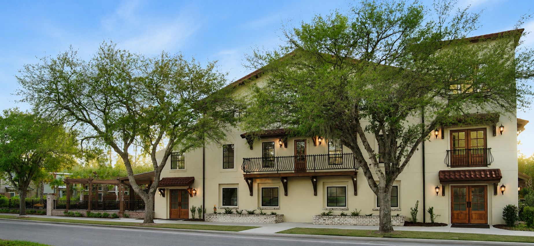 A multi-story residential building with white walls, wooden double doors, balconies with black railing, and exterior wall-mounted lantern lights. Large green trees with sprawling branches are in front of the building, and a sidewalk runs along the st