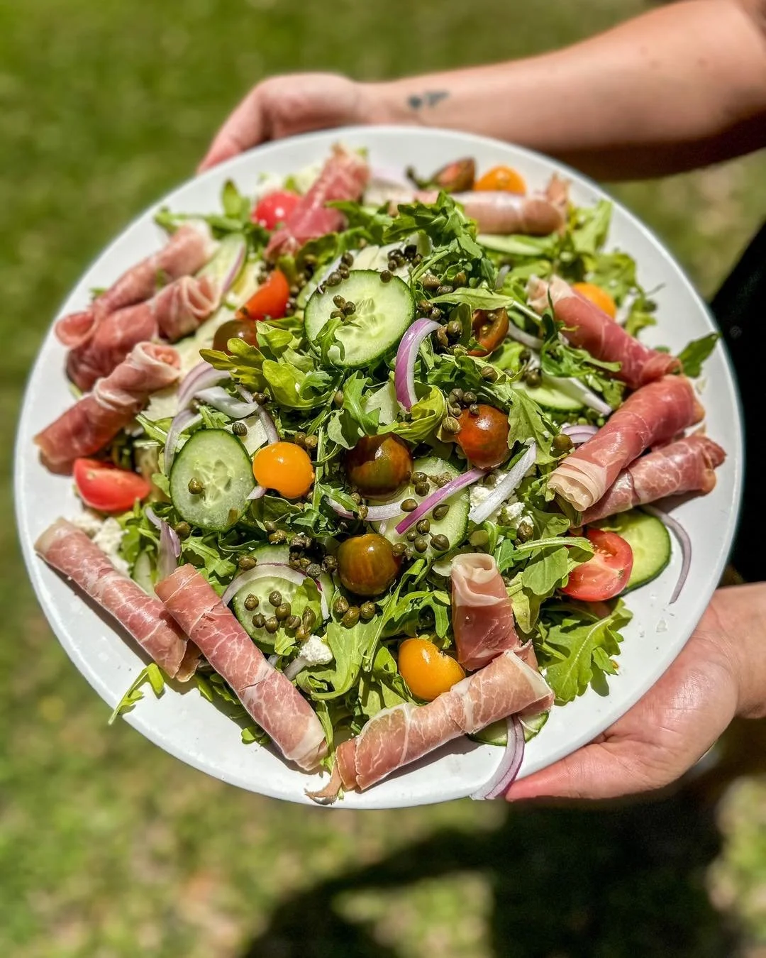 A person holding a large white plate filled with a mixed green salad topped with cherry tomatoes, cucumber slices, red onion, and black peppercorns, with rolled slices of prosciutto or similar cured meat around the perimeter.