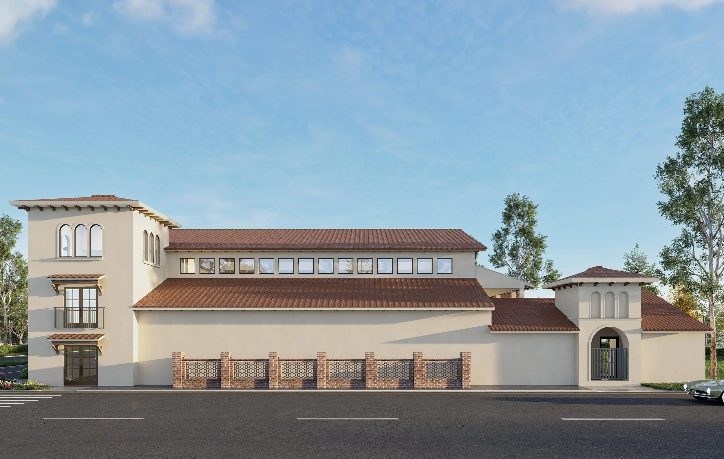 A large, modern building with a stucco exterior, red tile roof, and a combination of arched and rectangular windows. There is a decorative brick fence in front and a gated entrance to the right. The building is surrounded by trees and situated next to a road with a car on the right side.