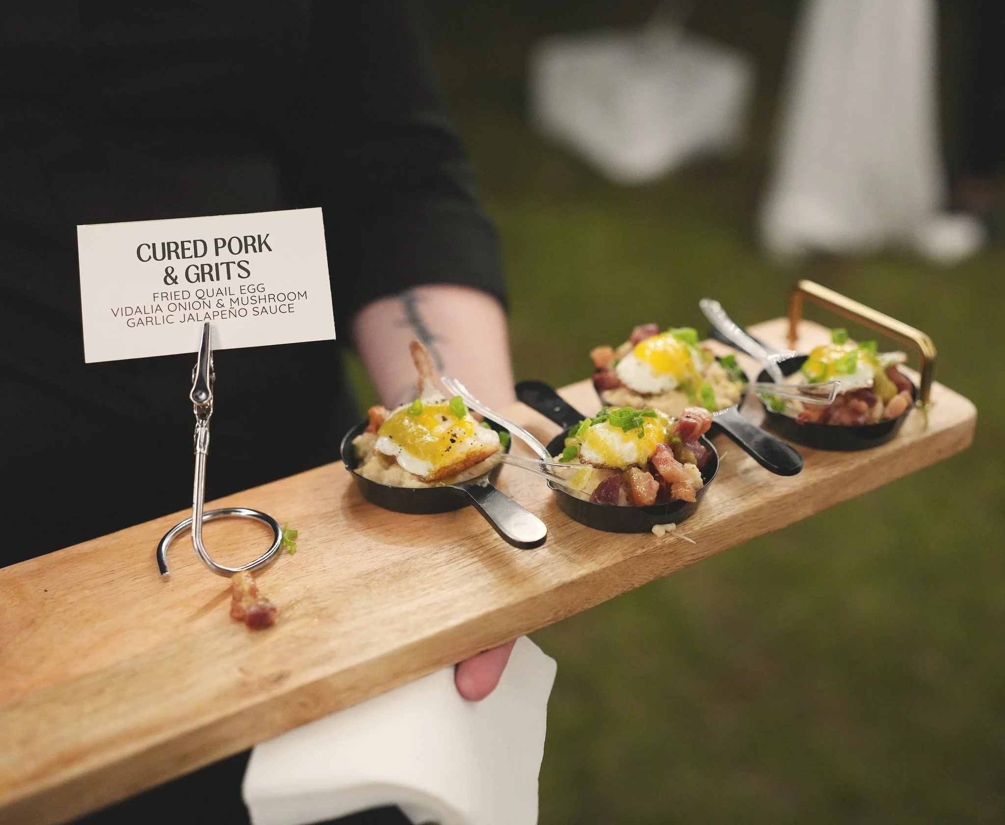 A person holding a wooden serving board with four small black cast iron skillets of food, topped with eggs and green garnish, labeled as 'CURED PORK & GRITS' with a description of fried quail egg, vidalia onion, mushroom, garlic, jalapeño sauce.