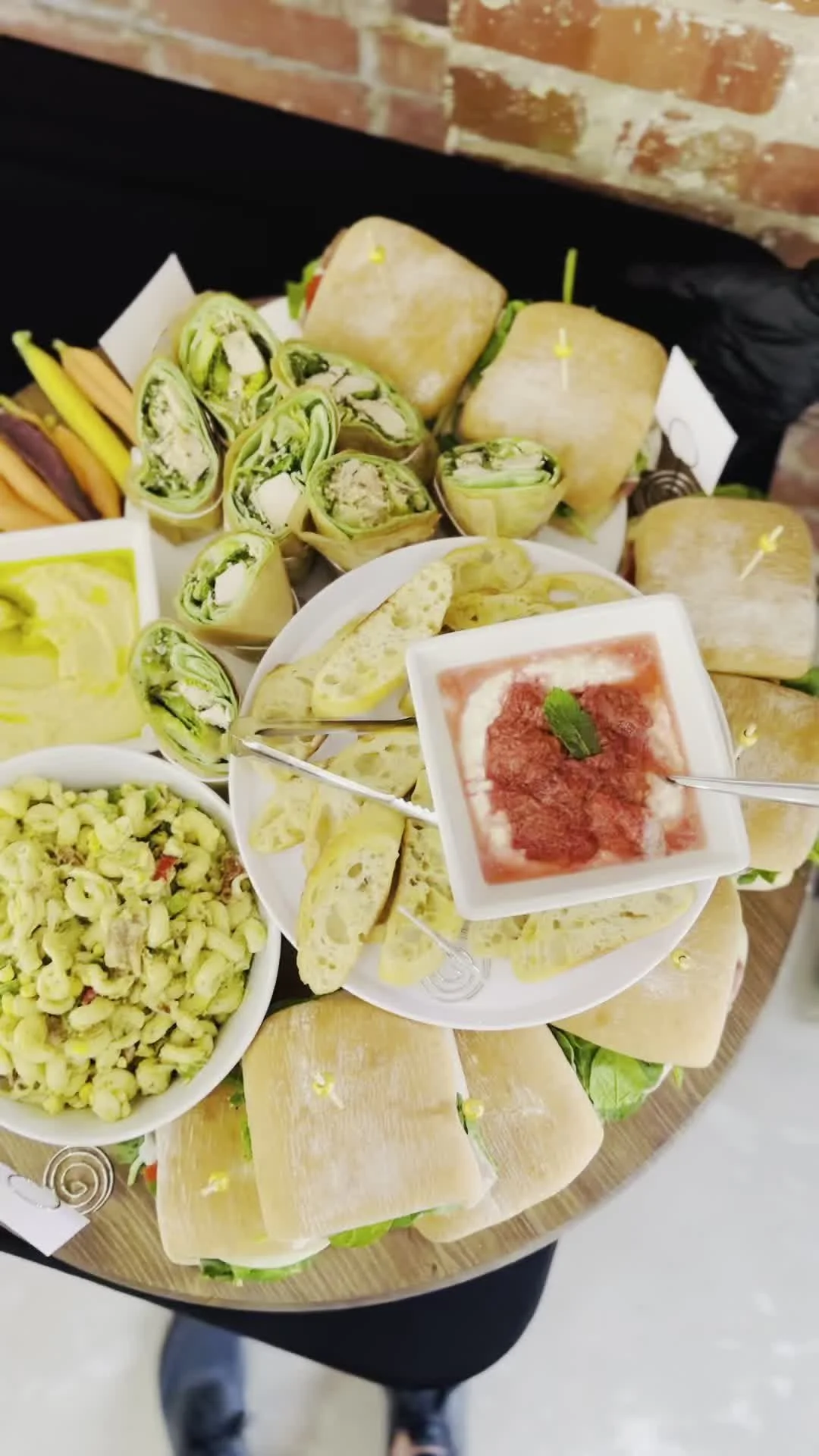 A round wooden platter with assorted wraps, sliced vegetables, a bowl of green pasta salad, and small dishes of sauces or dips