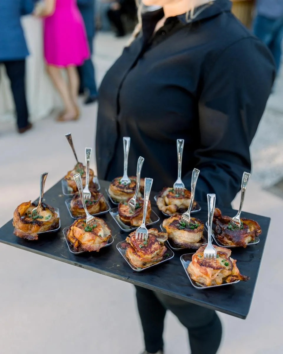 A server holds a tray with baked appetizers topped with herbs and served with forks, next to a person wearing a black shirt at an outdoor event.