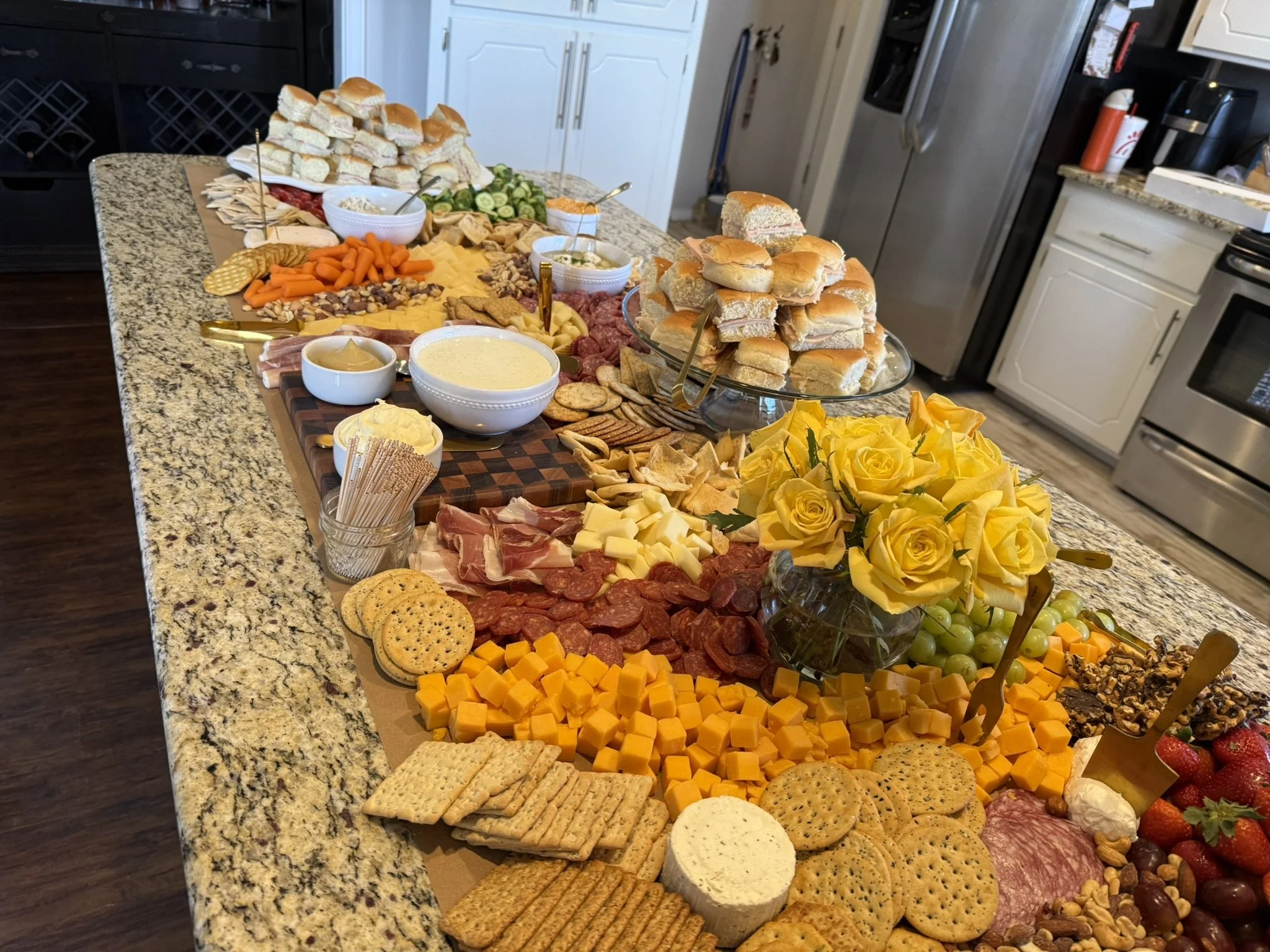 A large spread of sandwiches, cheese, meats, crackers, fruits, vegetables, and appetizers on a kitchen island for a party or gathering.