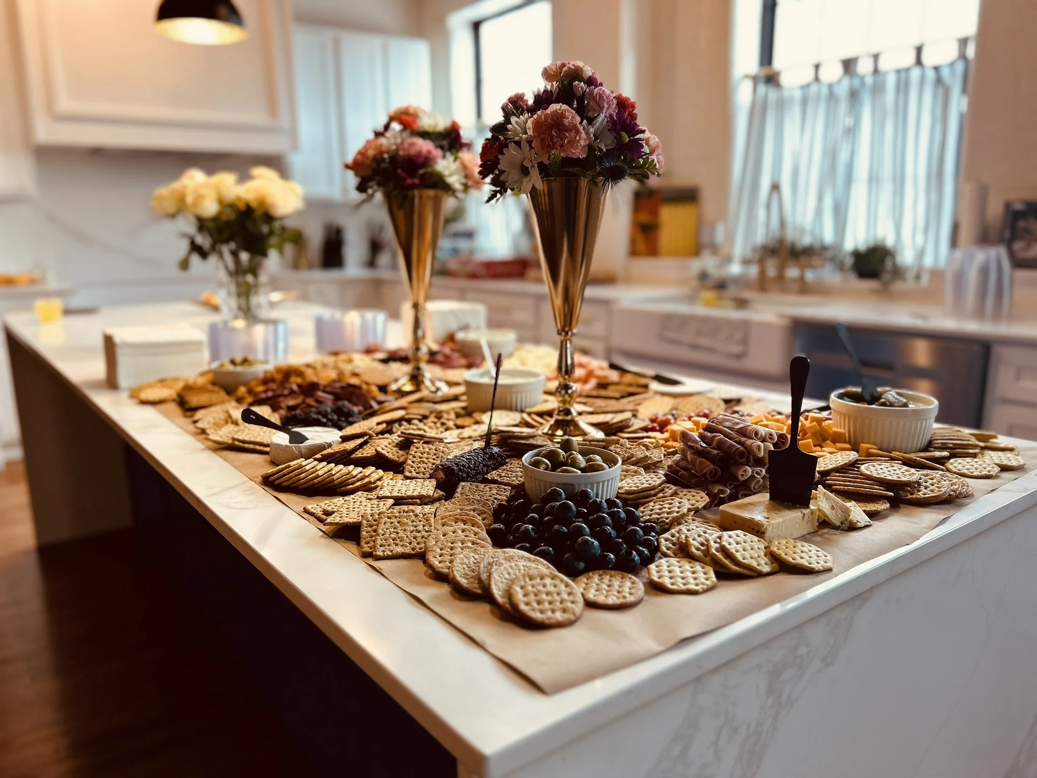 A cheese and cracker platter with bowls of olives and spread, decorated with tall floral arrangements on a kitchen island.