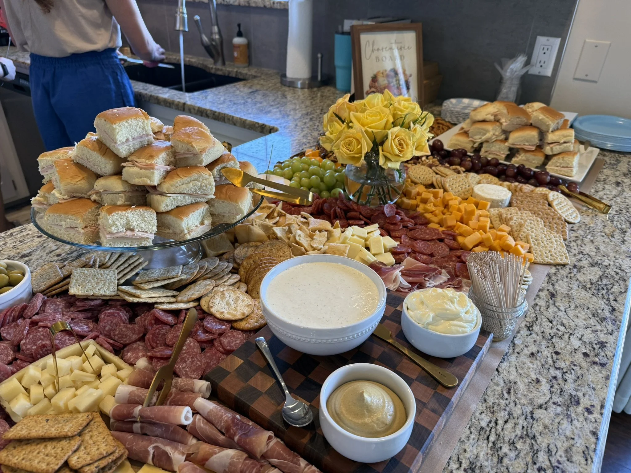 A table set with a charcuterie and sandwich platter, including cheese, meats, crackers, and grapes, accompanied by a bowl of dip and a bouquet of yellow roses. In the background, a person is washing dishes in a kitchen.