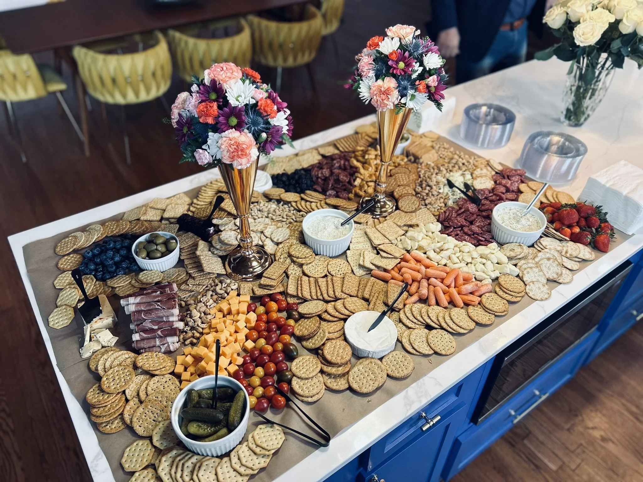 A charcuterie board with a variety of cheeses, meats, crackers, grapes, strawberries, cherry tomatoes, pickles, and snack bowls, decorated with three floral centerpieces in tall gold vases.