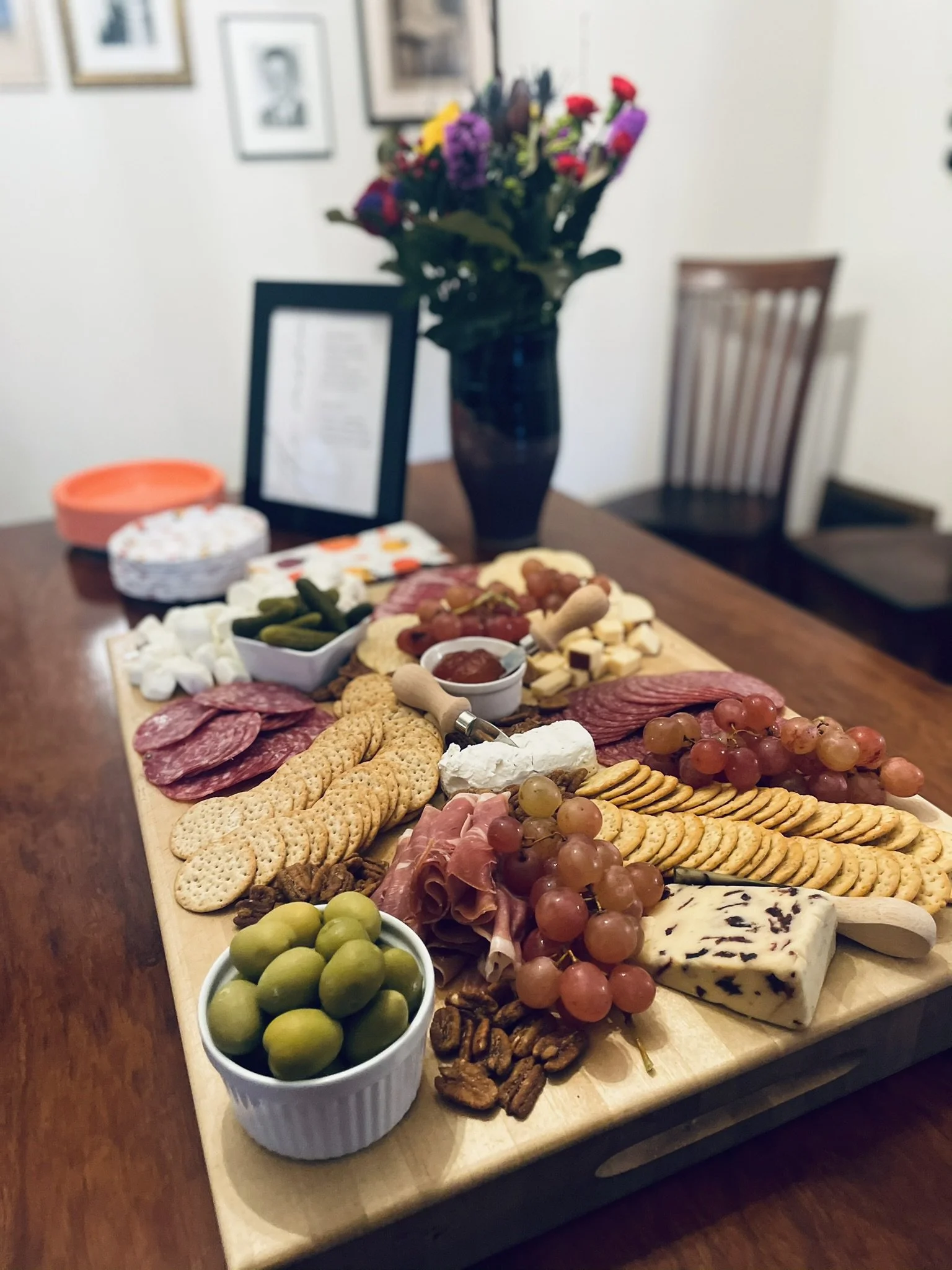 A charcuterie board with mixed cheeses, meats, grapes, olives, crackers, and nuts on a dining table, with a vase of colorful flowers in the background.
