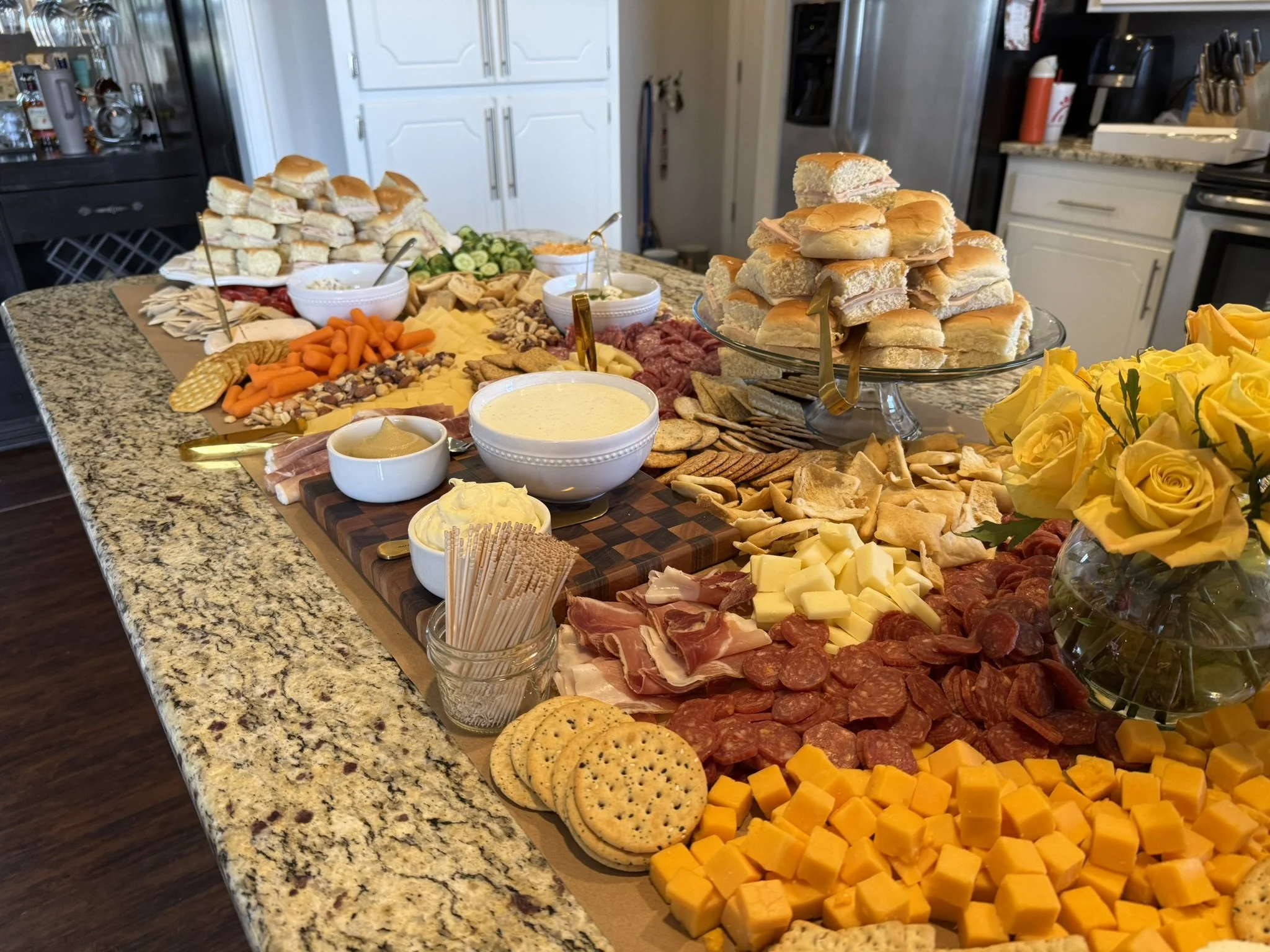 A large spread of finger foods on a kitchen island. Includes sandwiches, cheese, deli meats, crackers, vegetables, dips, and a vase of yellow roses.