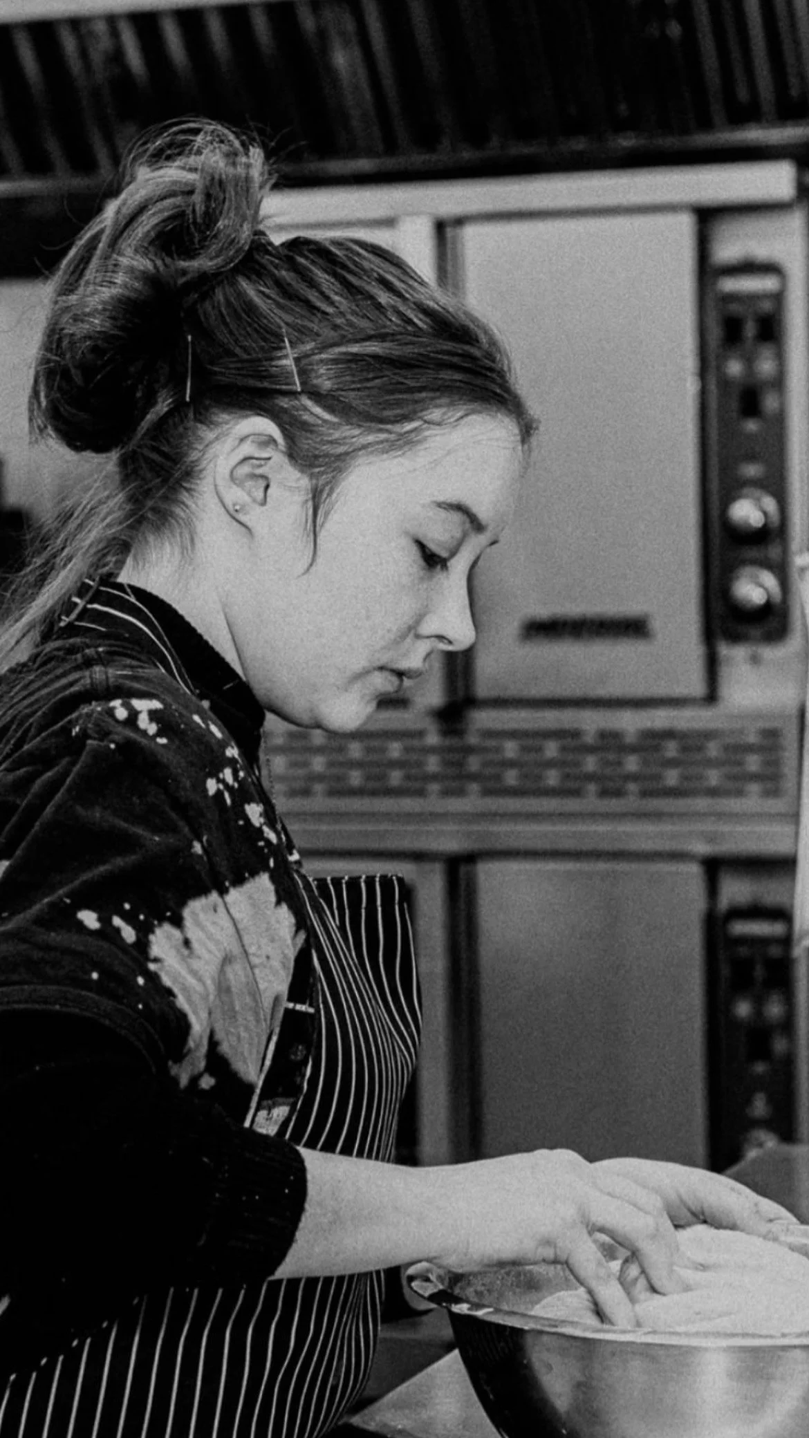 A woman with long hair tied up, wearing an apron, is working with dough in a bowl in a kitchen setting.
