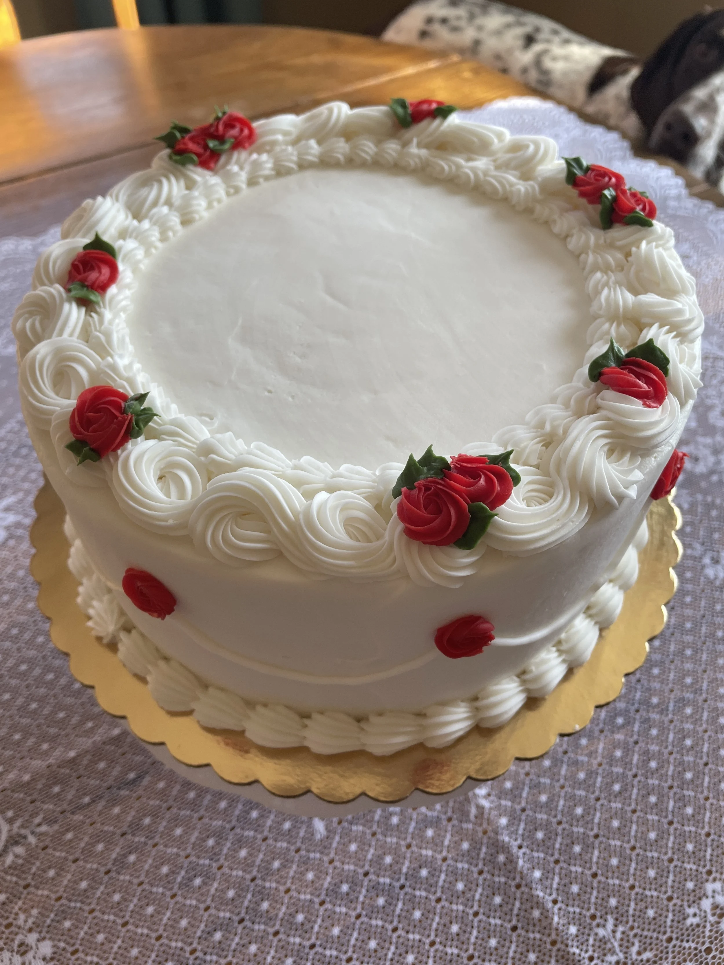 Round white cake decorated with red and green icing roses around the top edge, sitting on a gold cake board on a lace tablecloth.