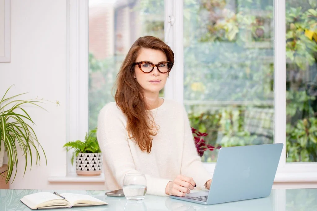 Woman with glasses working on a laptop at a glass-top table in a bright room with large window and plants.