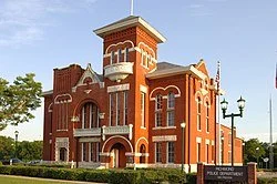 A red brick historic building with a tower and arched windows, situated on a street corner with a flagpole.