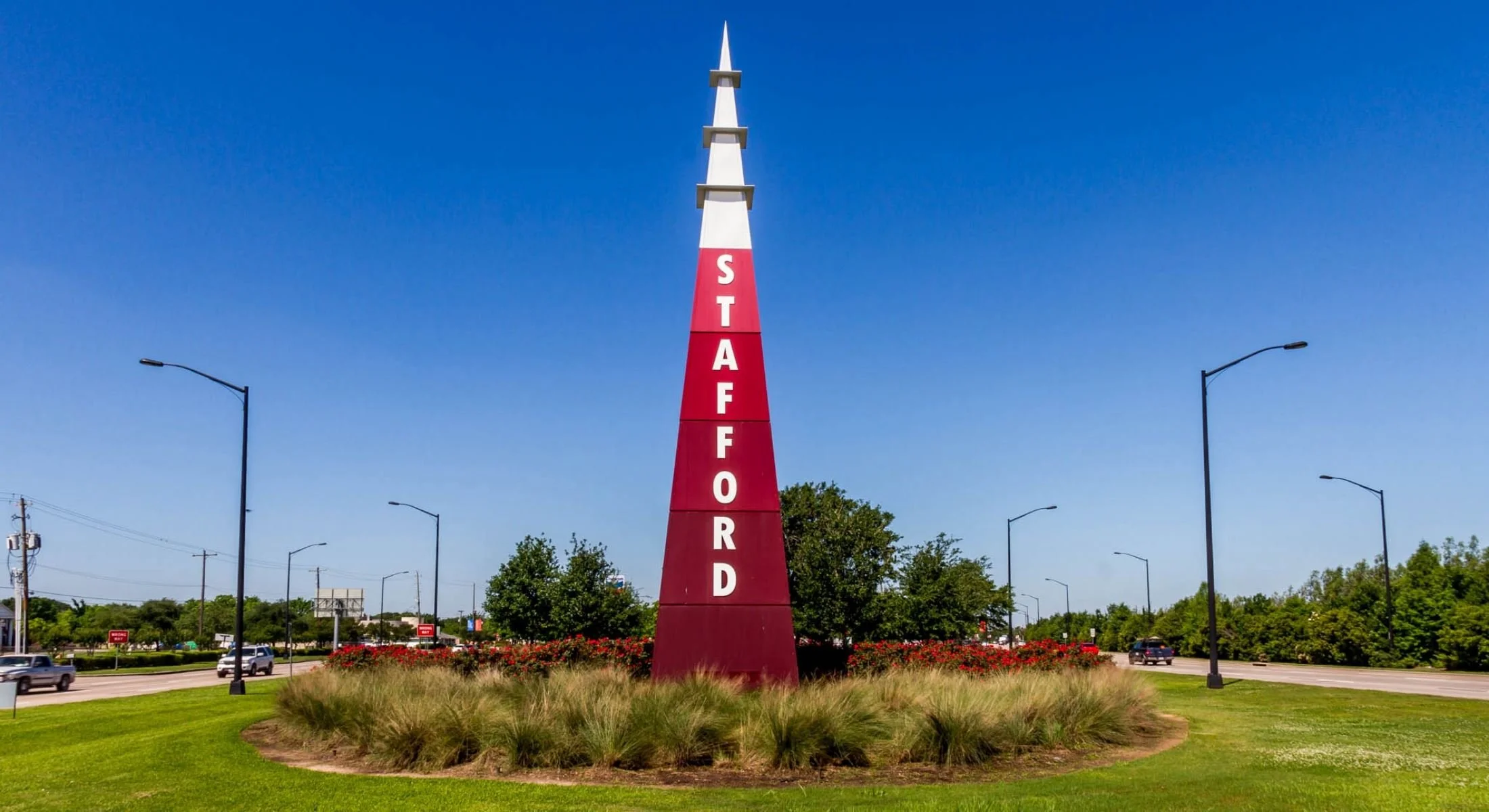 A tall, red and white directional sign with the word 'STAFFORD' on it, situated in a landscaped roundabout with grass, bushes, and trees, under a clear blue sky.