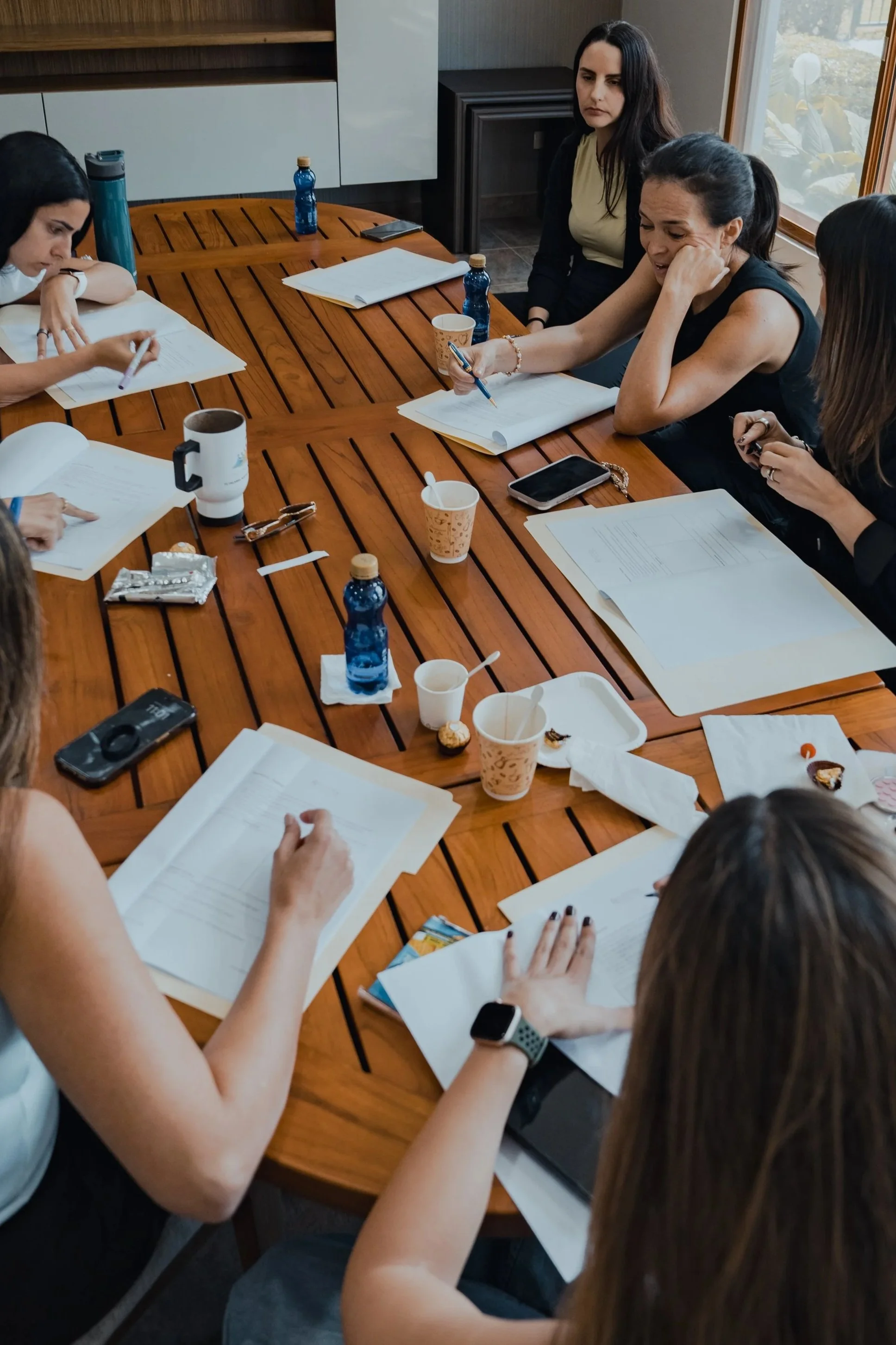 Grupo de mujeres en una reunión de trabajo alrededor de una mesa de madera con documentos, bebidas y snacks.