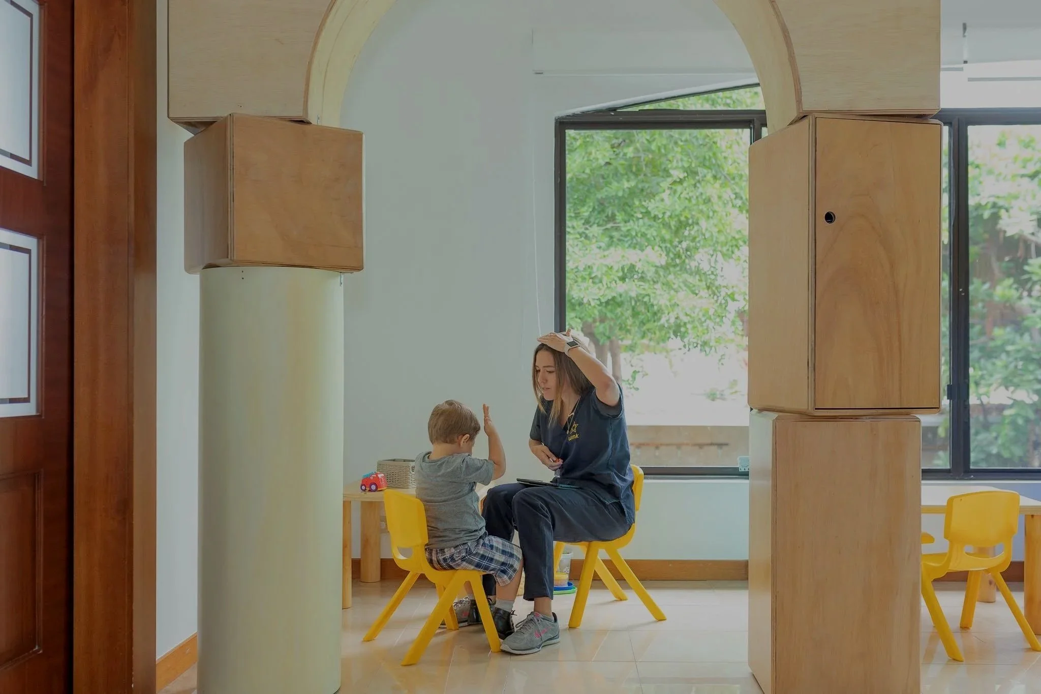Una mujer y un niño sentado en sillas amarillas en un aula o sala de juegos, interactuando. La mujer está usando guantes y parece estar en una actividad de enseñanza o cuidado, con una ventana grande al fondo que muestra árboles verdes.