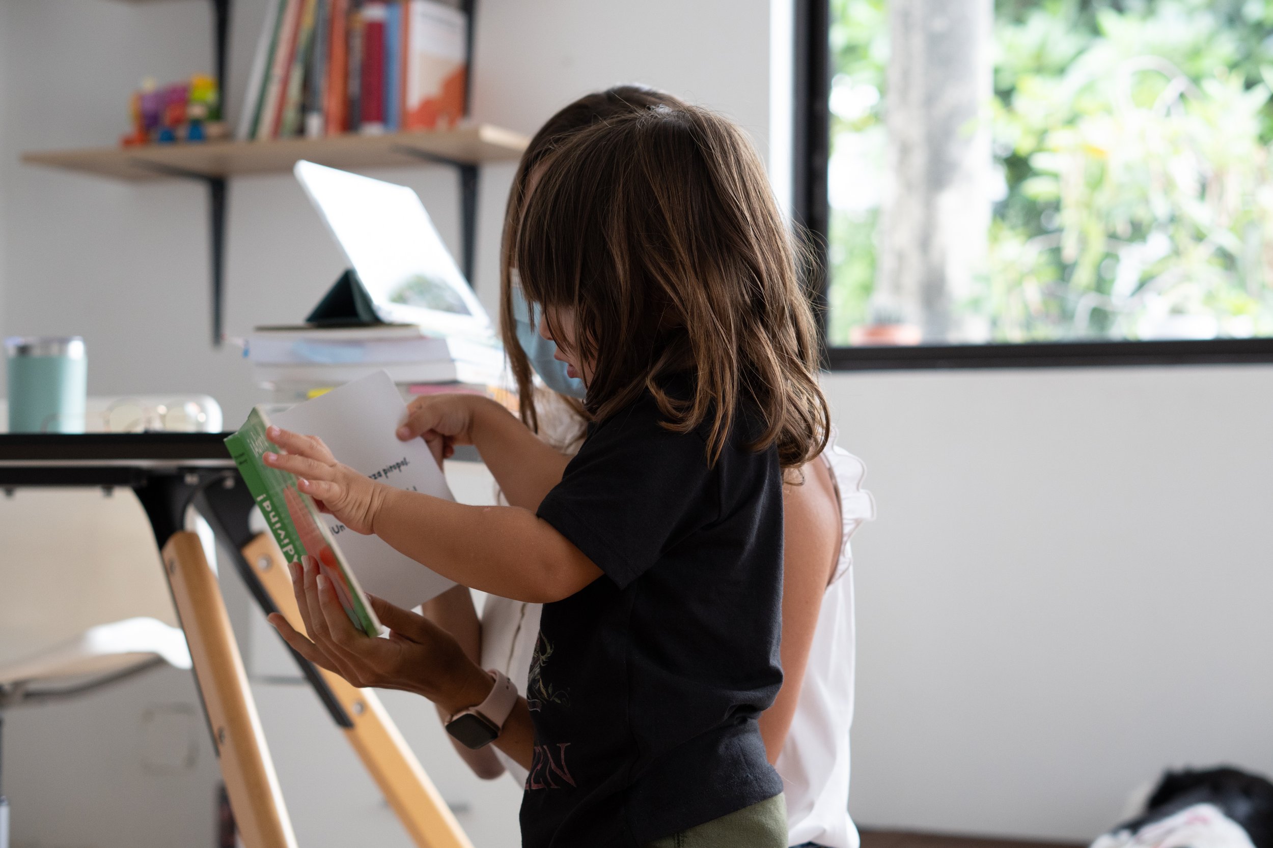 Niña con mascarilla leyendo un libro en una habitación con escritorio y estantes con libros.