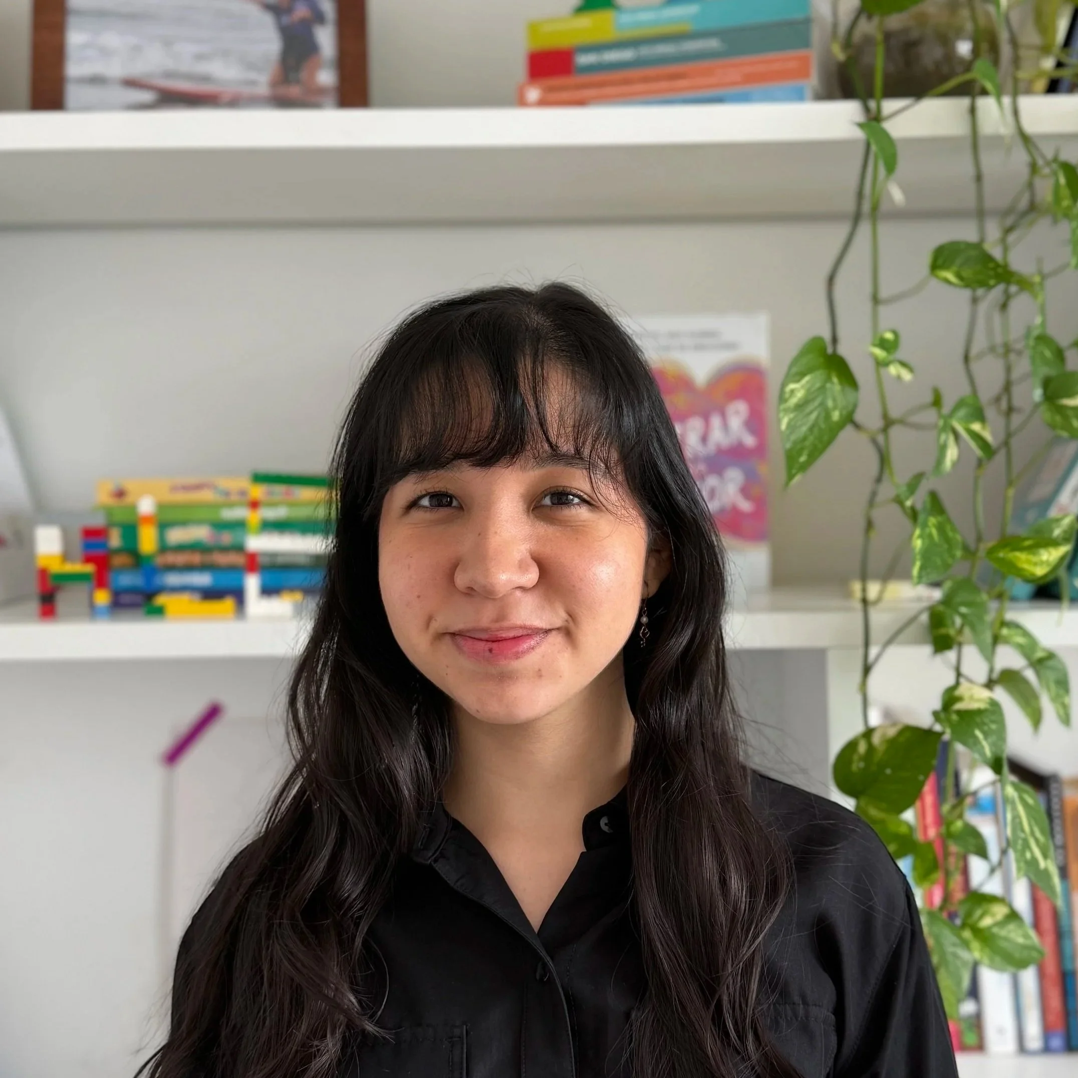 Joven mujer con cabello largo y oscuro, sonriendo, en un fondo de estantería con libros y plantas.