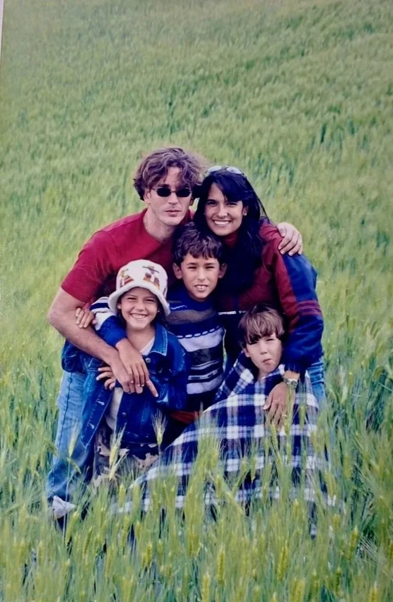 Una familia de cinco personas en un campo de césped. Dos adultos y tres niños sonriendo y abrazándose.