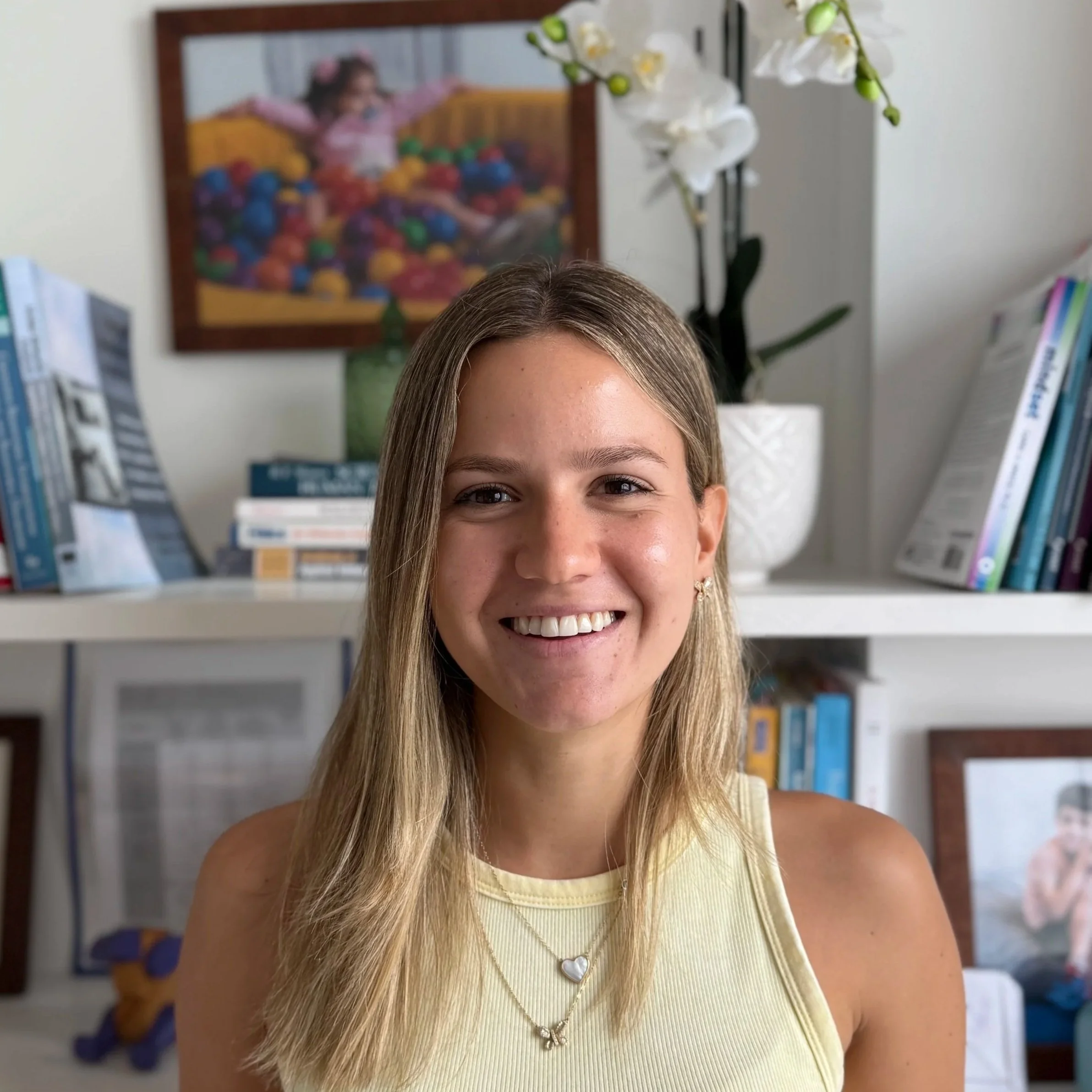 Retrato de una joven sonriendo, con fondo de libreros, plantas y cuadros en una habitación.