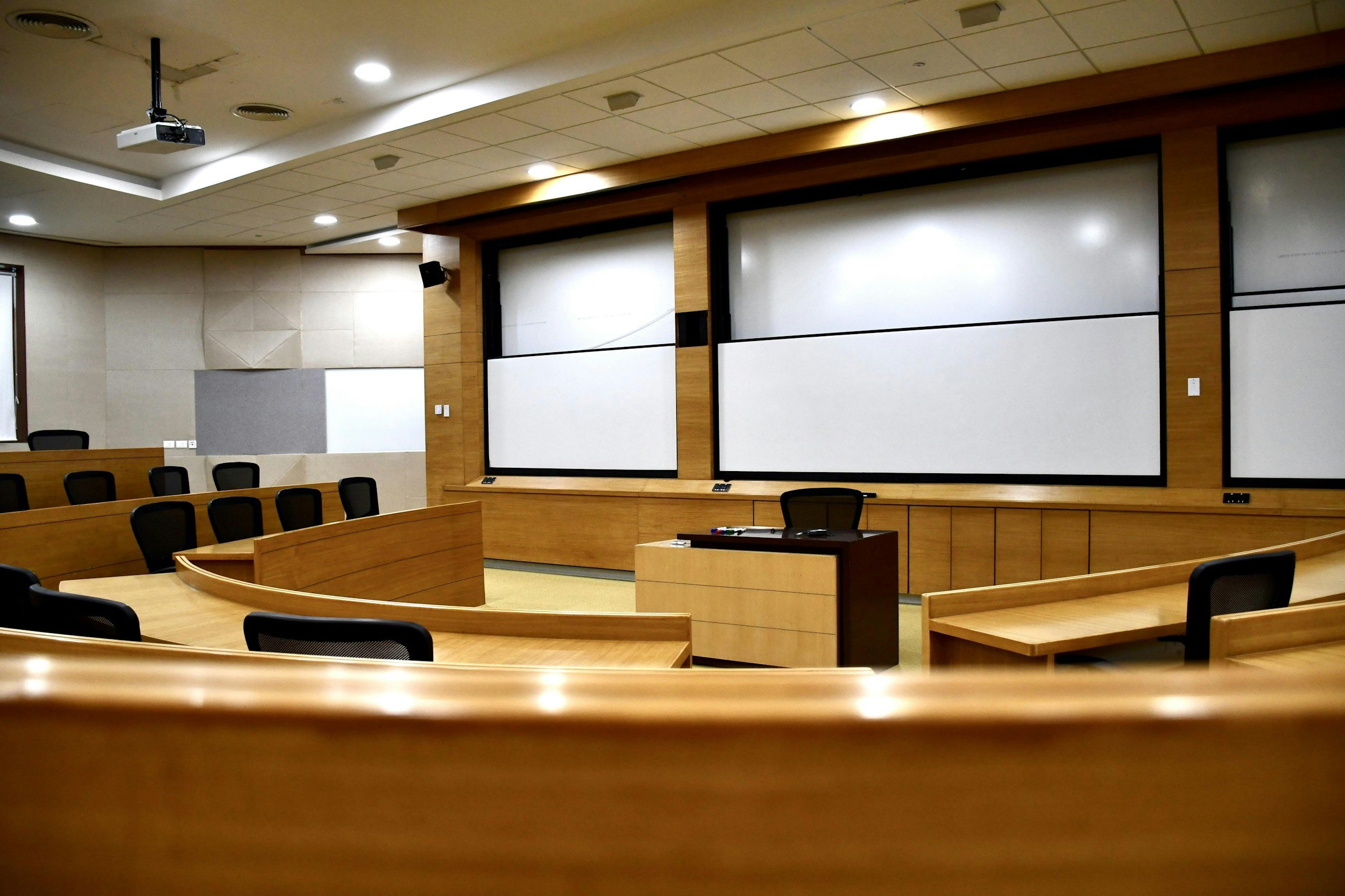 A modern conference room with wooden desks, black chairs, white projection screens, and ceiling lights.