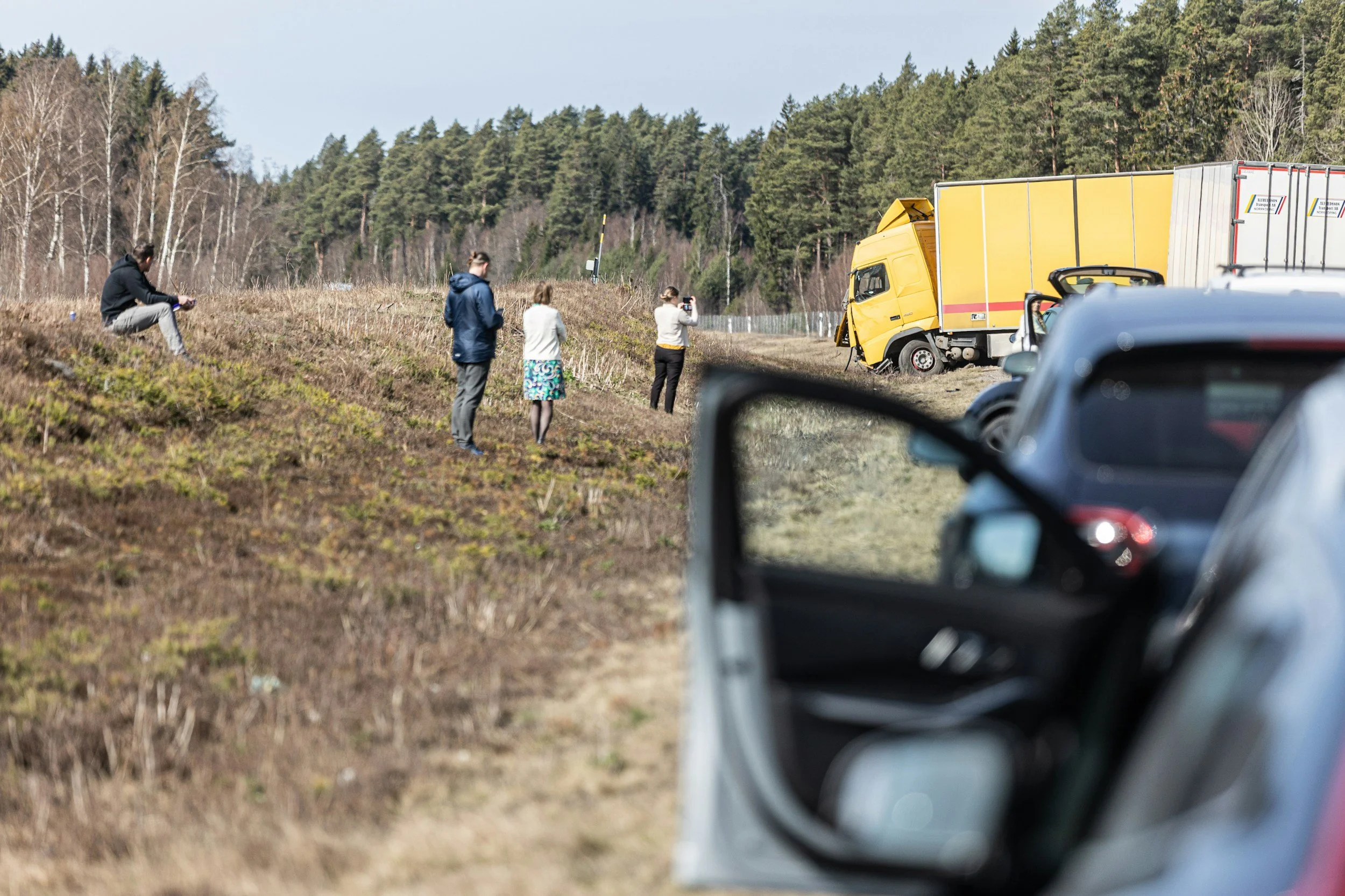 People taking photos of a yellow truck crash on the side of the road in a rural area surrounded by trees.