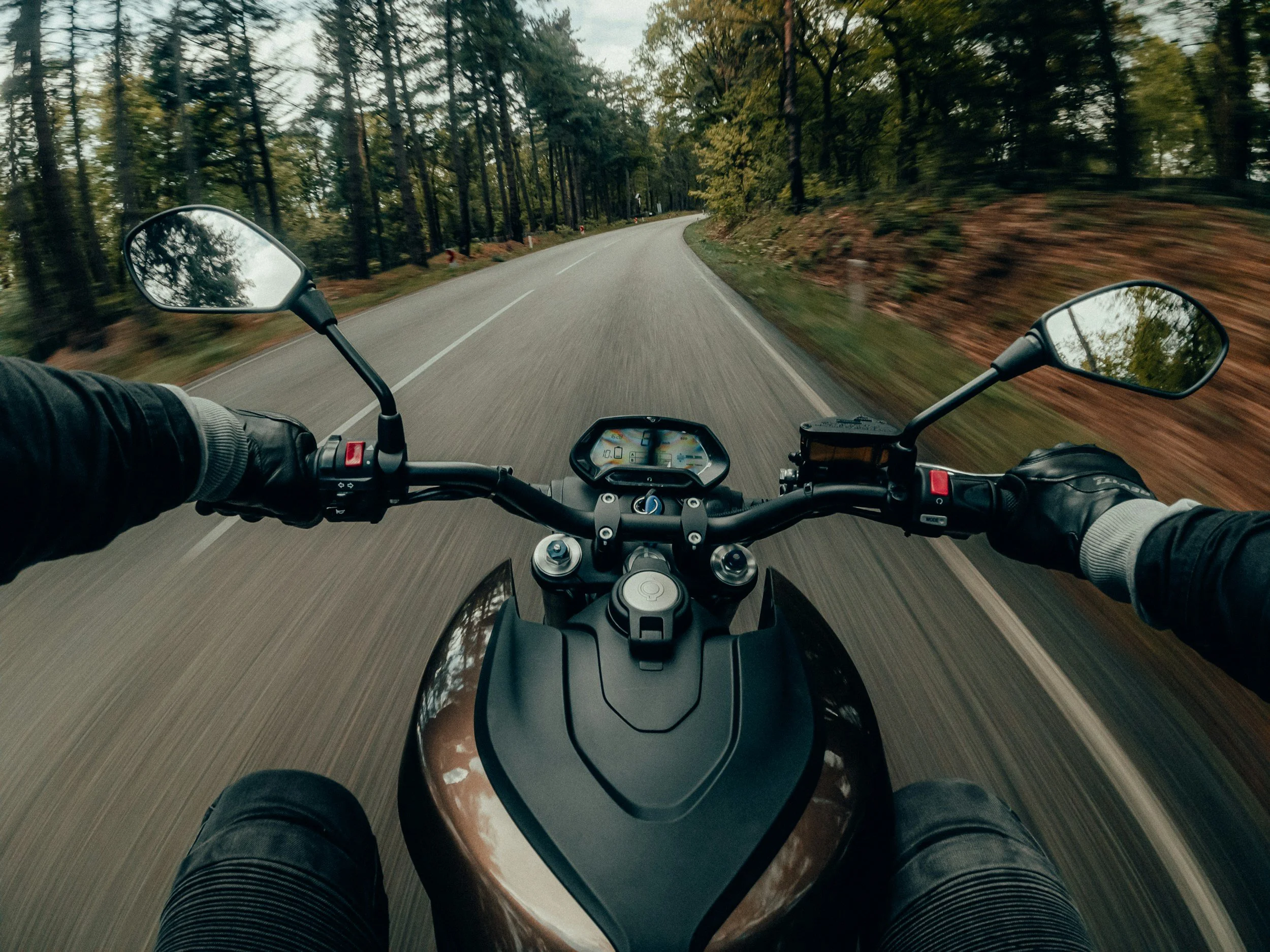 First-person view of a motorcycle rider on a curving, tree-lined road surrounded by green and yellow foliage.