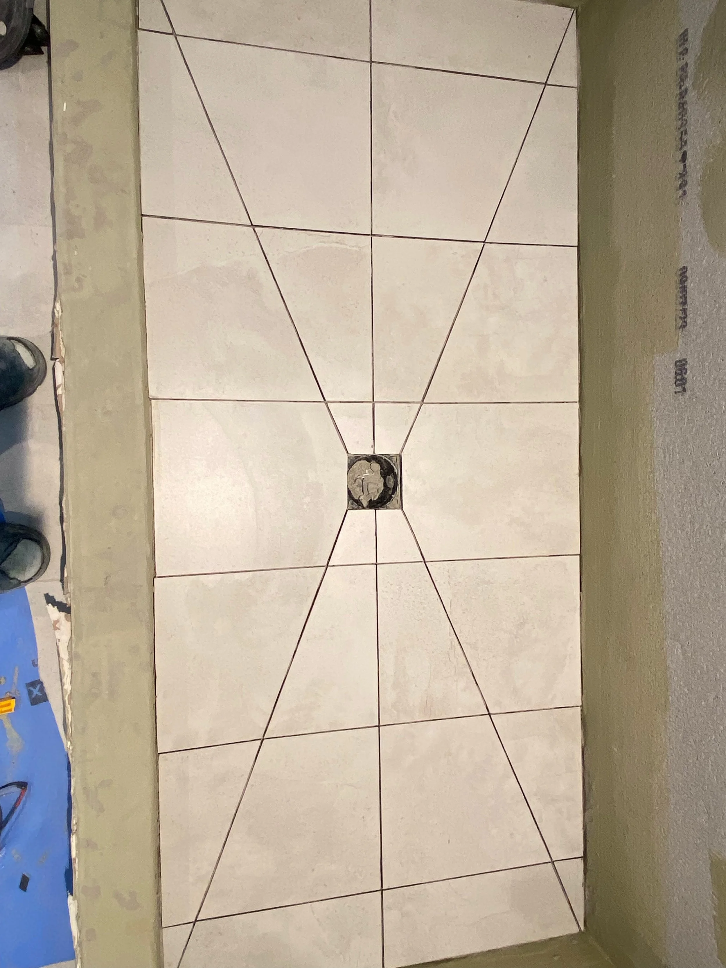 A section of floor with partially installed white tiles, some grout lines visible, and a central drain hole. Utility tools and parts are visible at the edges of the image.