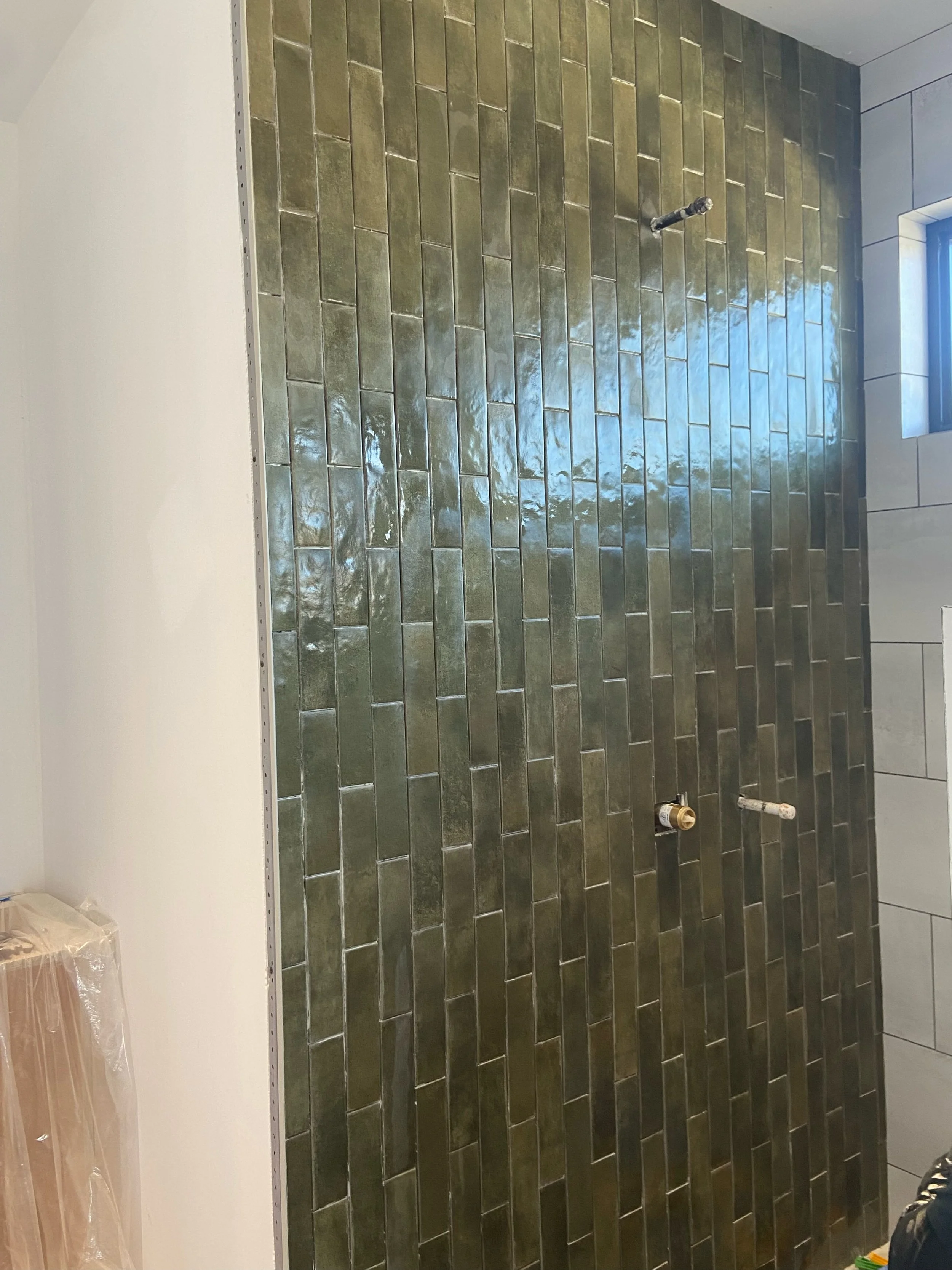 A bathroom shower area under construction with green rectangular tiles on the wall, chrome plumbing fixtures, and a window on the right side.