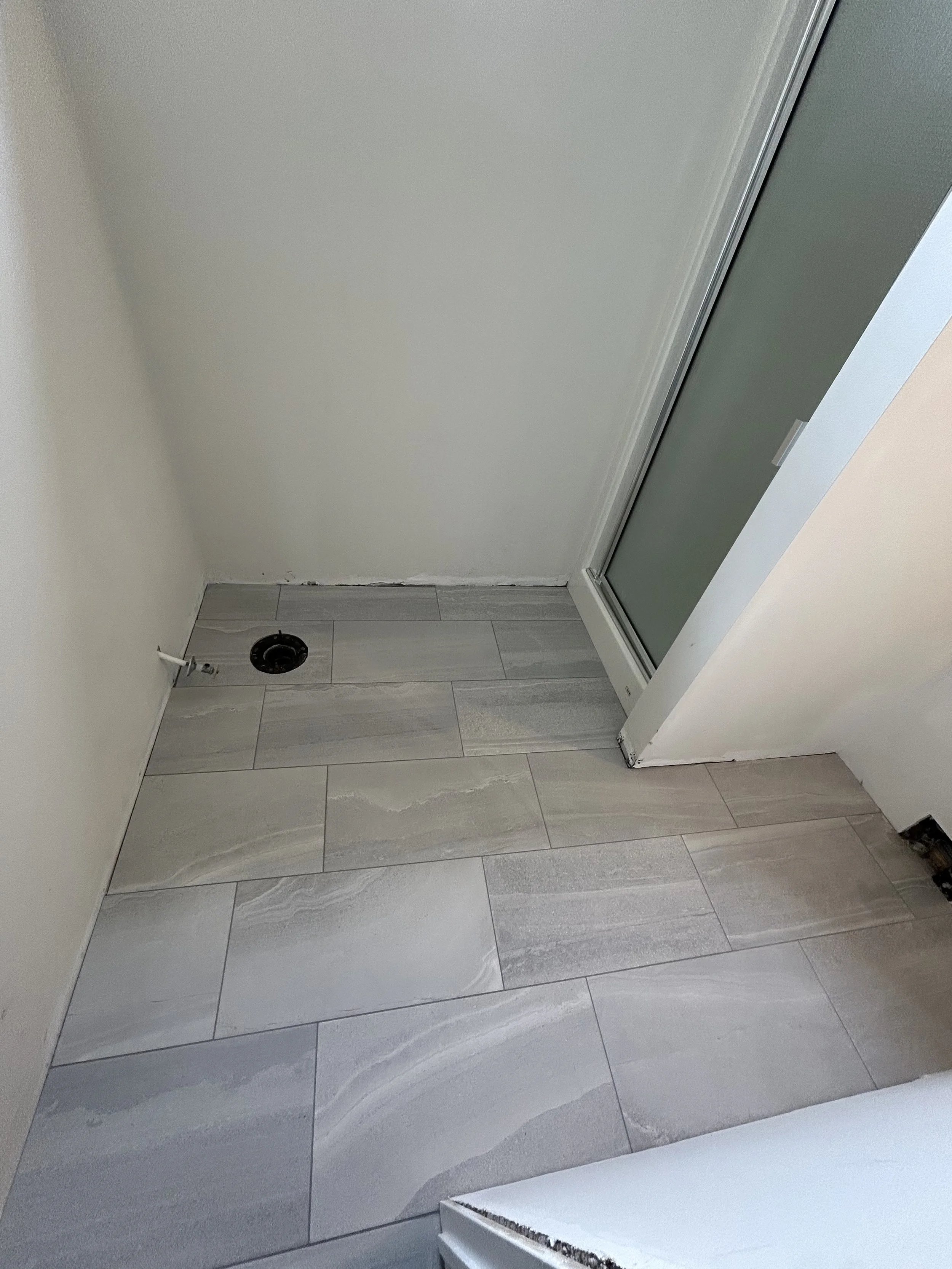 Corner of a bathroom or laundry room with gray tiled flooring, a recessed drain, and a sliding glass door or shower enclosure.