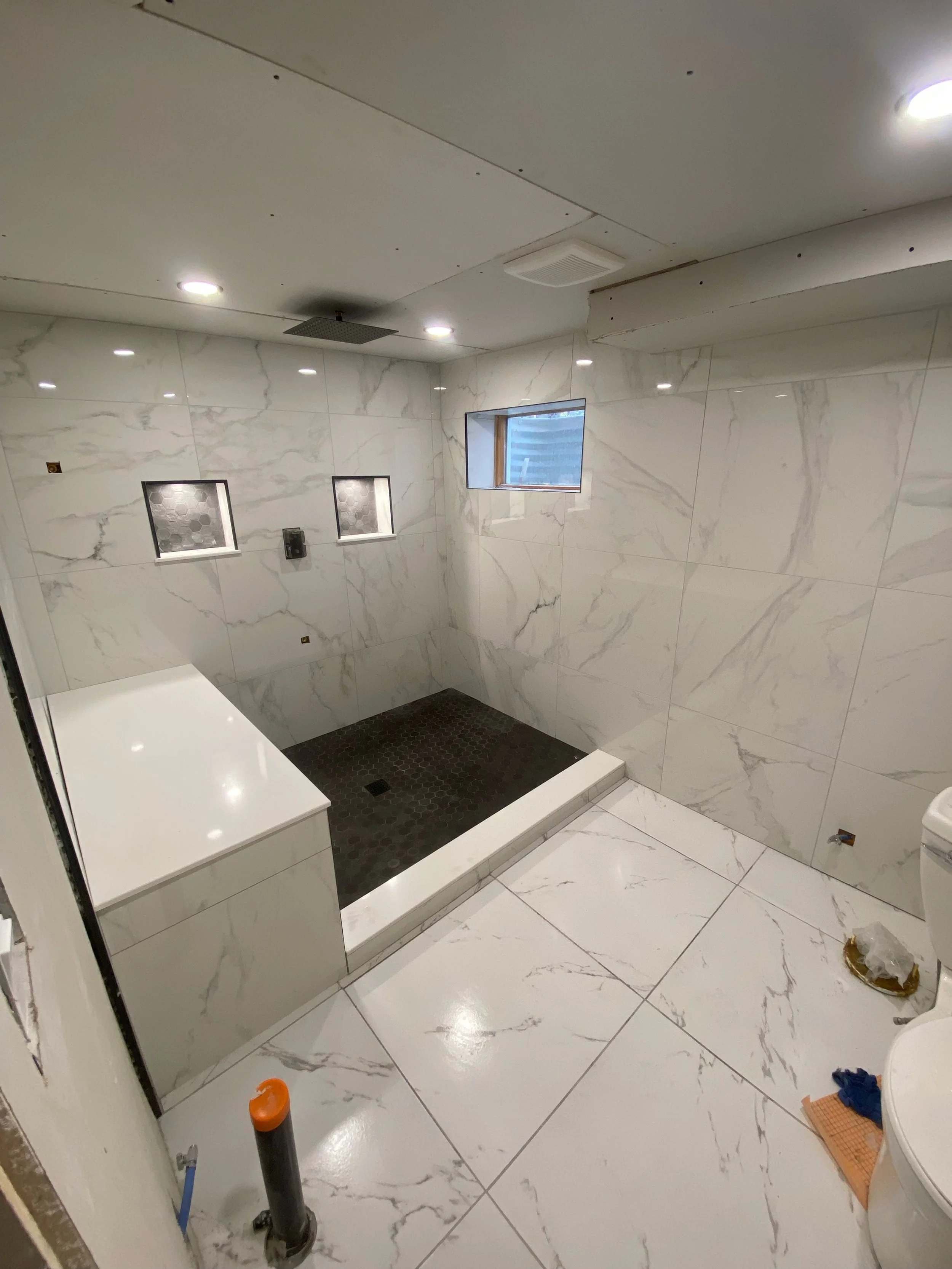 Bathroom under construction with marble tiles, a shower area with black tiles, small windows, and construction materials on the floor.