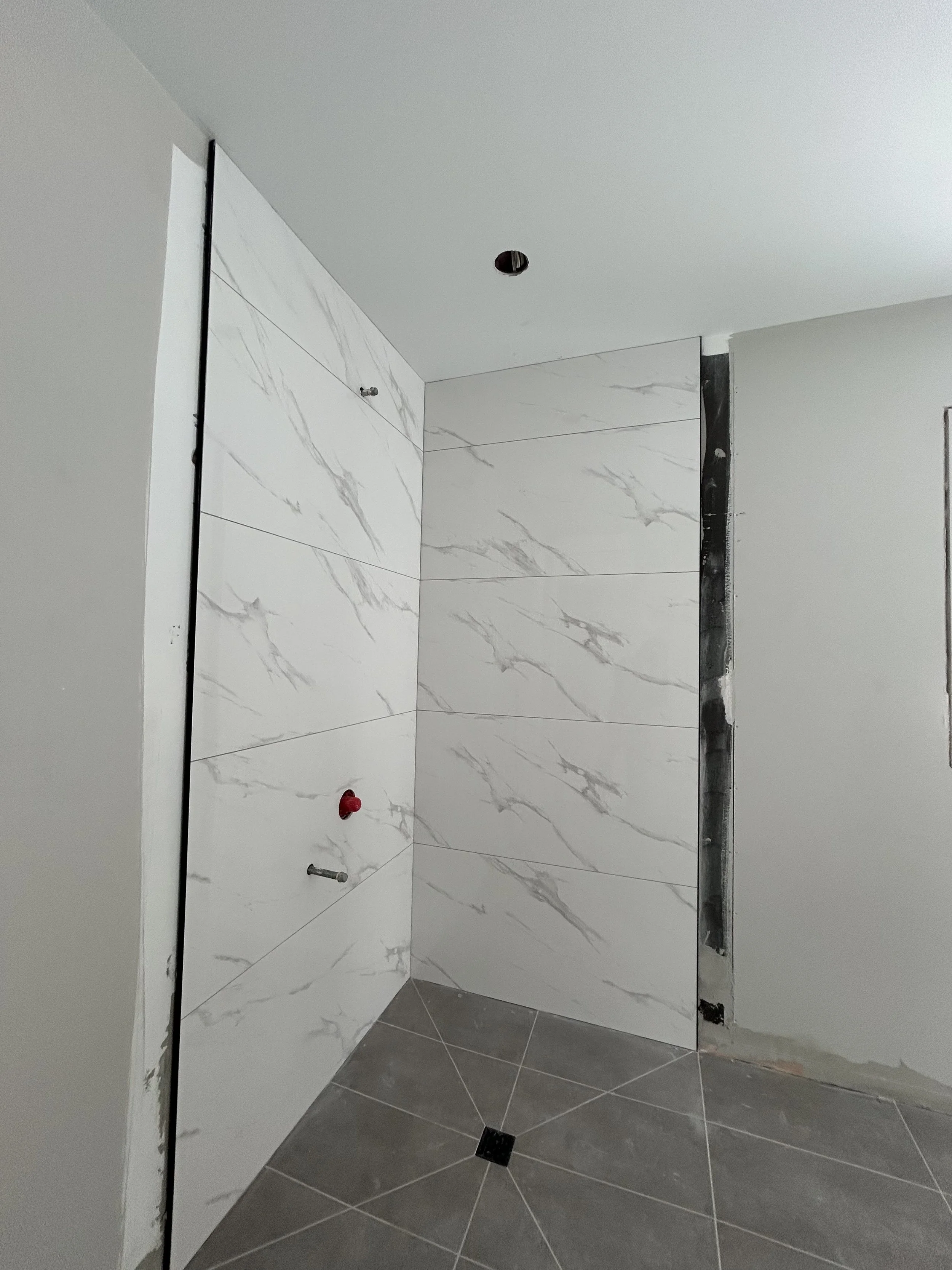 Interior of a bathroom under construction with white marble wall tiles, a floor drain, and an unfinished section near the door frame.