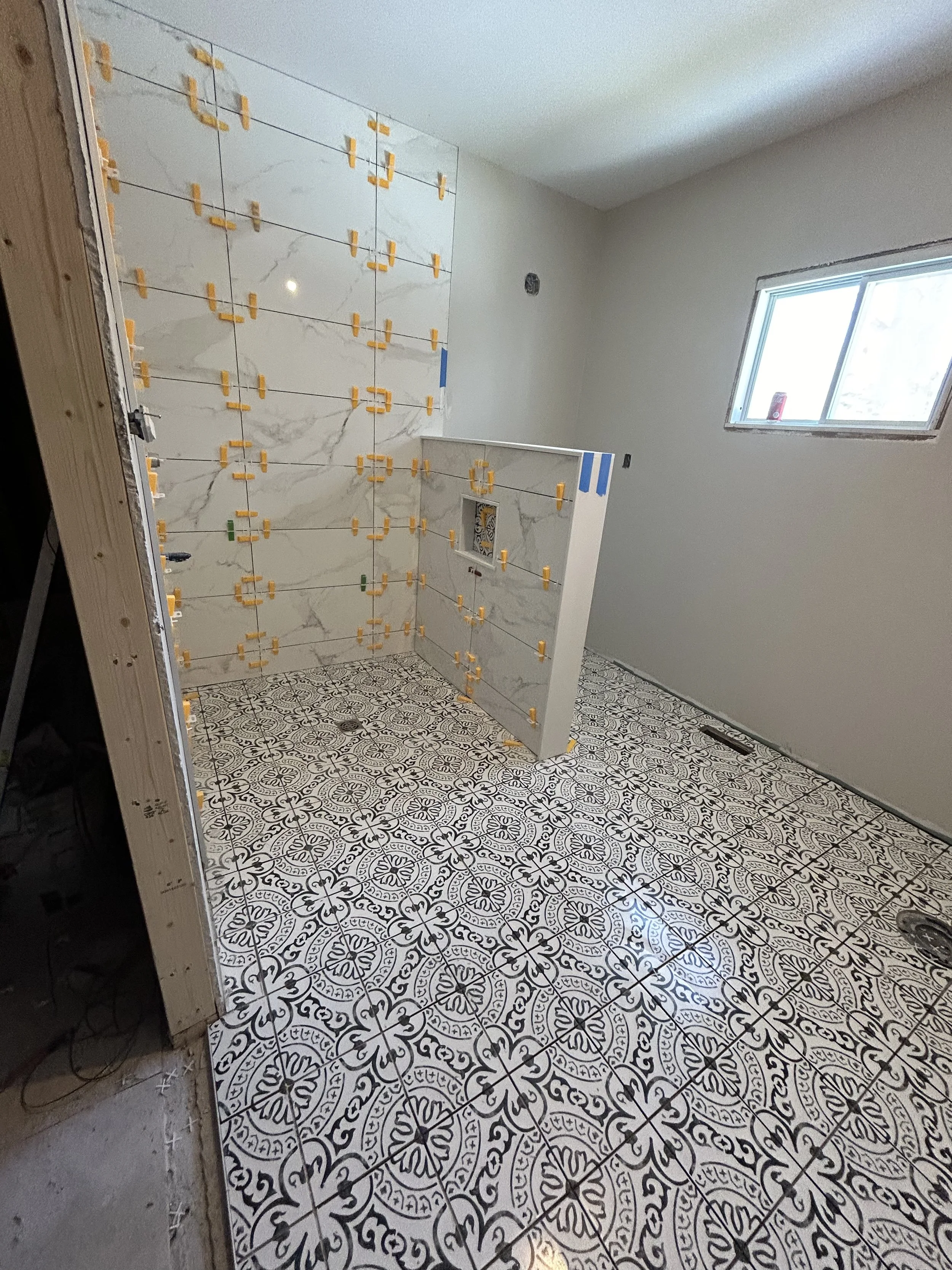 Bathroom under construction with decorative black and white patterned floor tiles, a partially tiled wall with large marble look tiles, and an opening for a shower or bathtub installation. Construction clips are visible on the wall tiles.