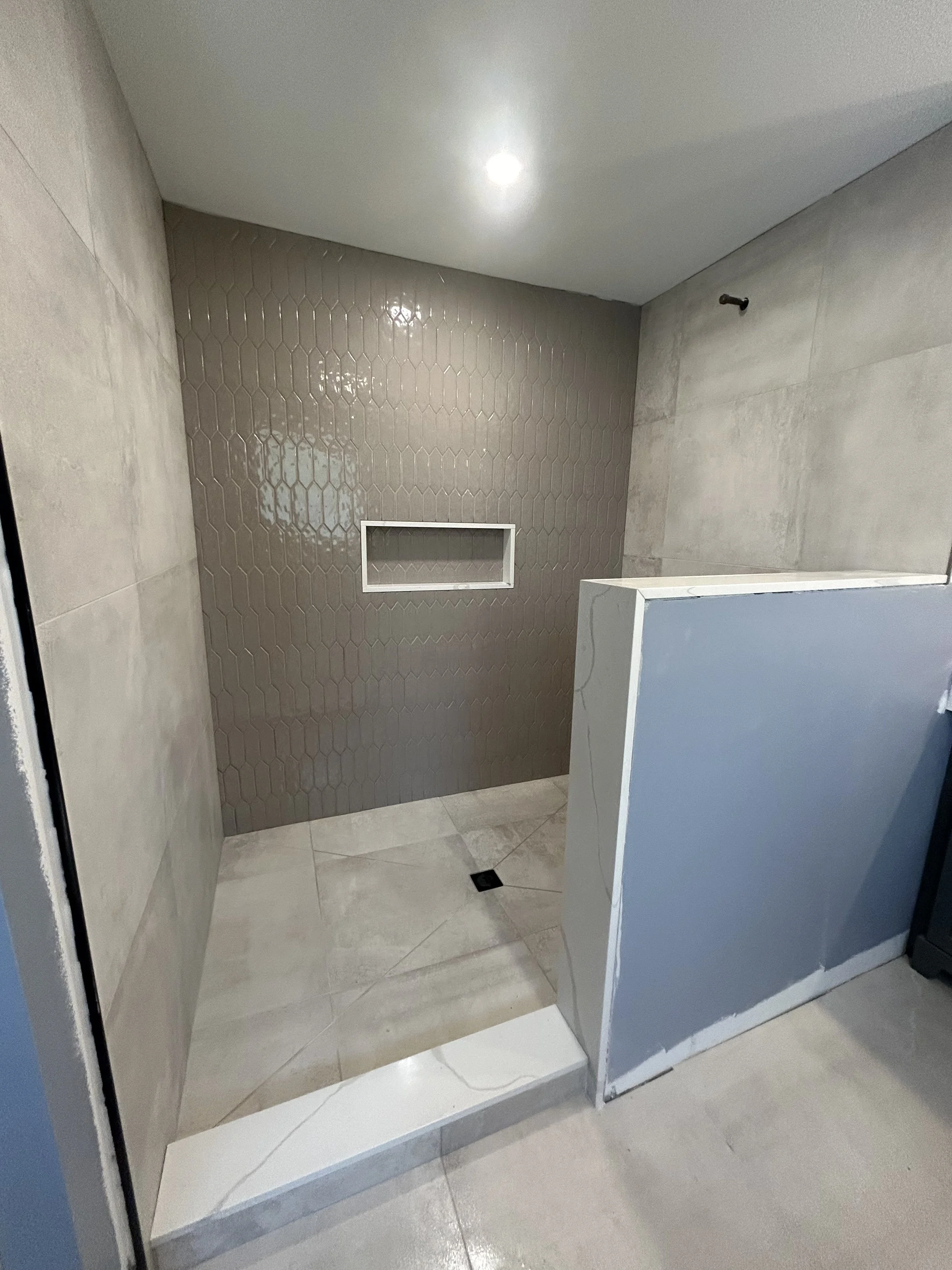 An unfinished shower area with beige and gray walls, a built-in niche, and a floor drain, with a partial wall and no shower fixtures installed.