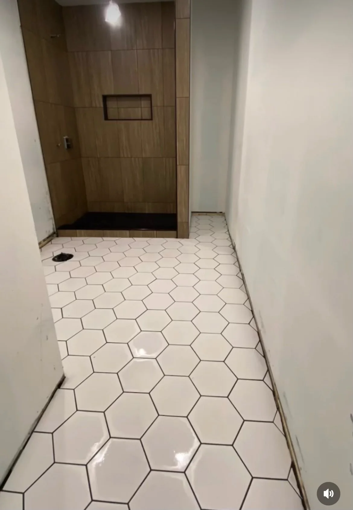 Empty bathroom with white hexagonal tile flooring, a wooden shower stall with a small built-in shelf, and plain white walls.
