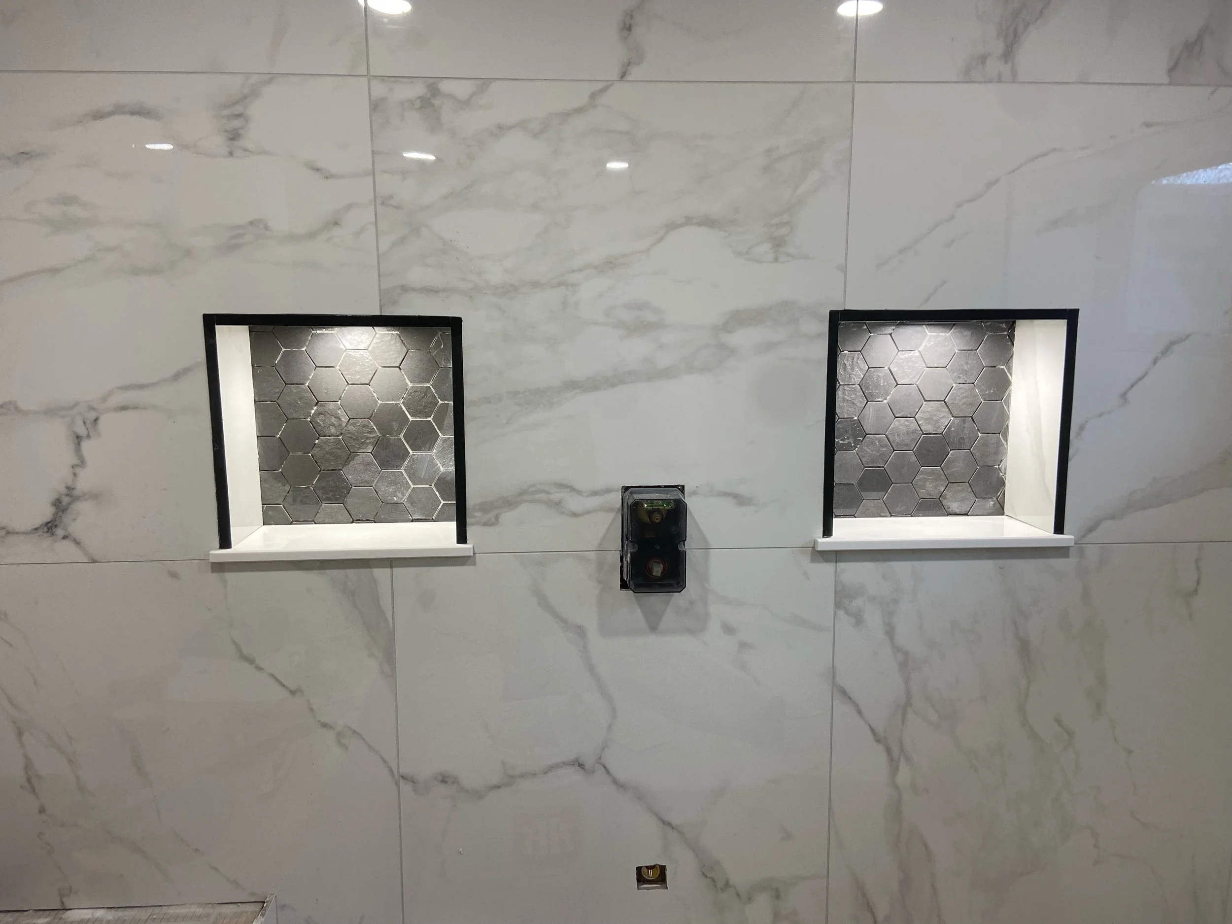Two built-in wall niches with black frames and gray hexagon tile backs on a white marble wall, with a central electrical box between them.