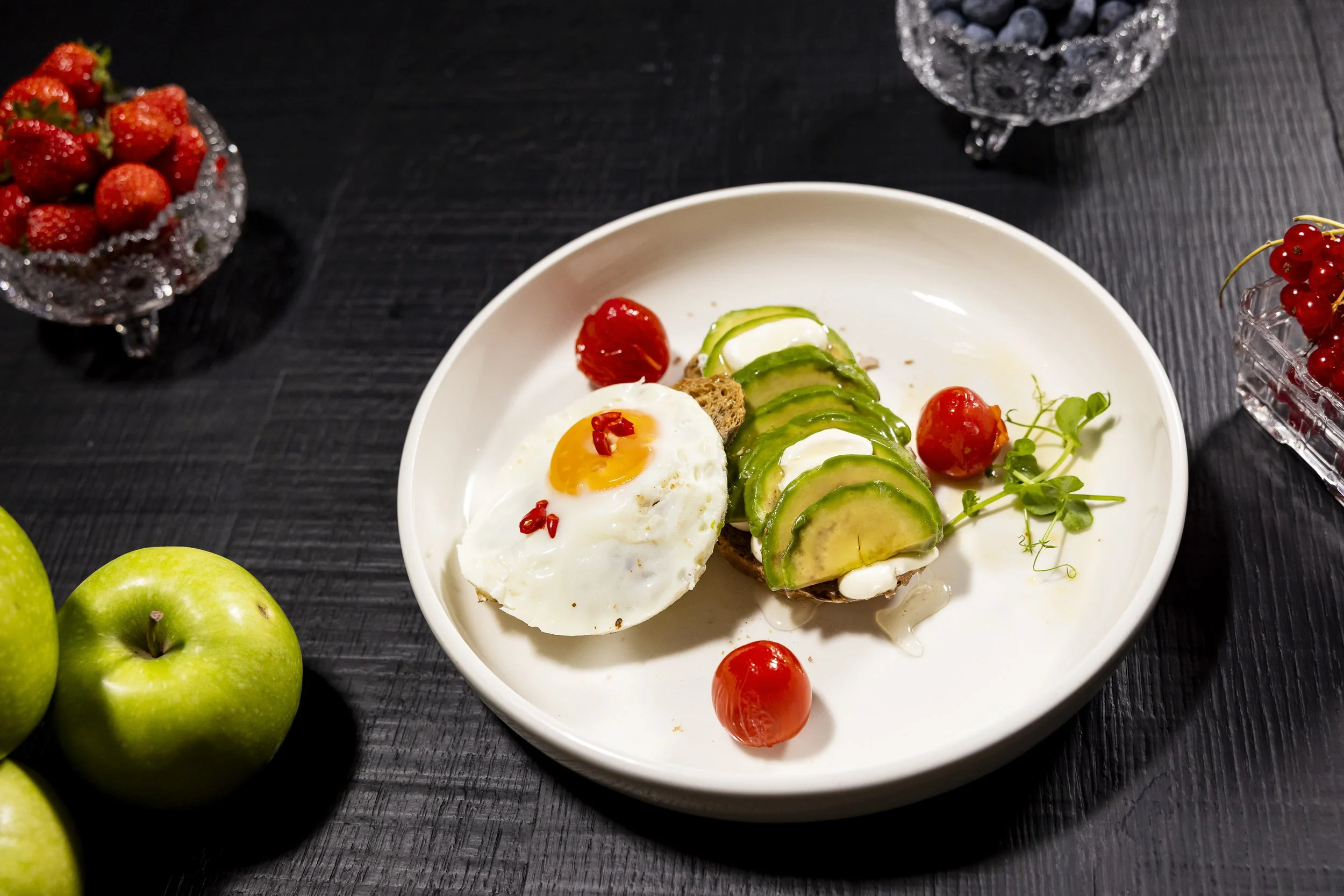 Uova in camicia con avocado, pomodorini e pane integrale su piatto bianco.