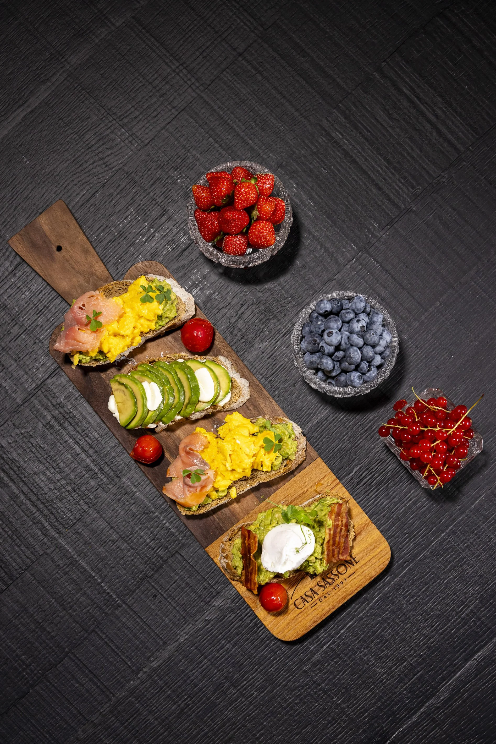 Una tavola con quattro open sandwiches e tre ciotole di frutta fresca. Le fette di pane sono condite con vari ingredienti come salmone, uova strapazzate, avocado, e cetrioli, decorati con erbe aromatiche.