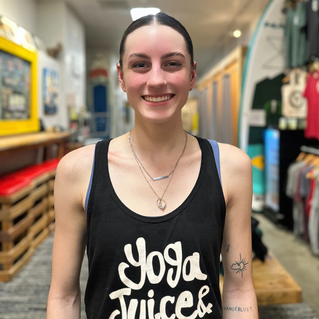 A smiling woman in a pink tank top with 'Yoga Juice & More' logo, standing in a gym or studio, with hands on hips, wearing a necklace and watch.