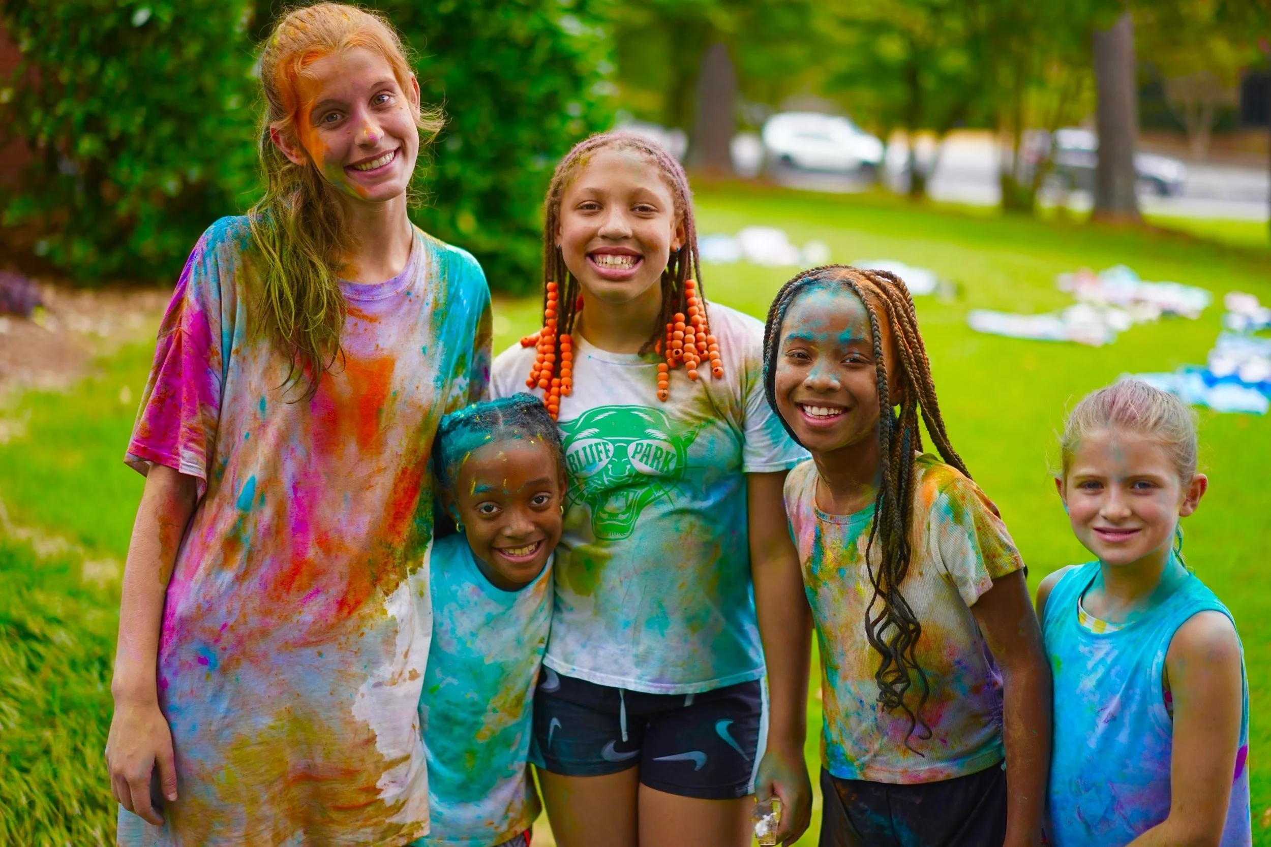Group of seven young girls at summer camp, all wearing matching beige t-shirts with a forest logo and the words 'Camp Adventure.' They have colorful volunteer lanyards with name tags around their necks and are smiling happily, standing in a hallway with a wooden door and an exit sign overhead.
