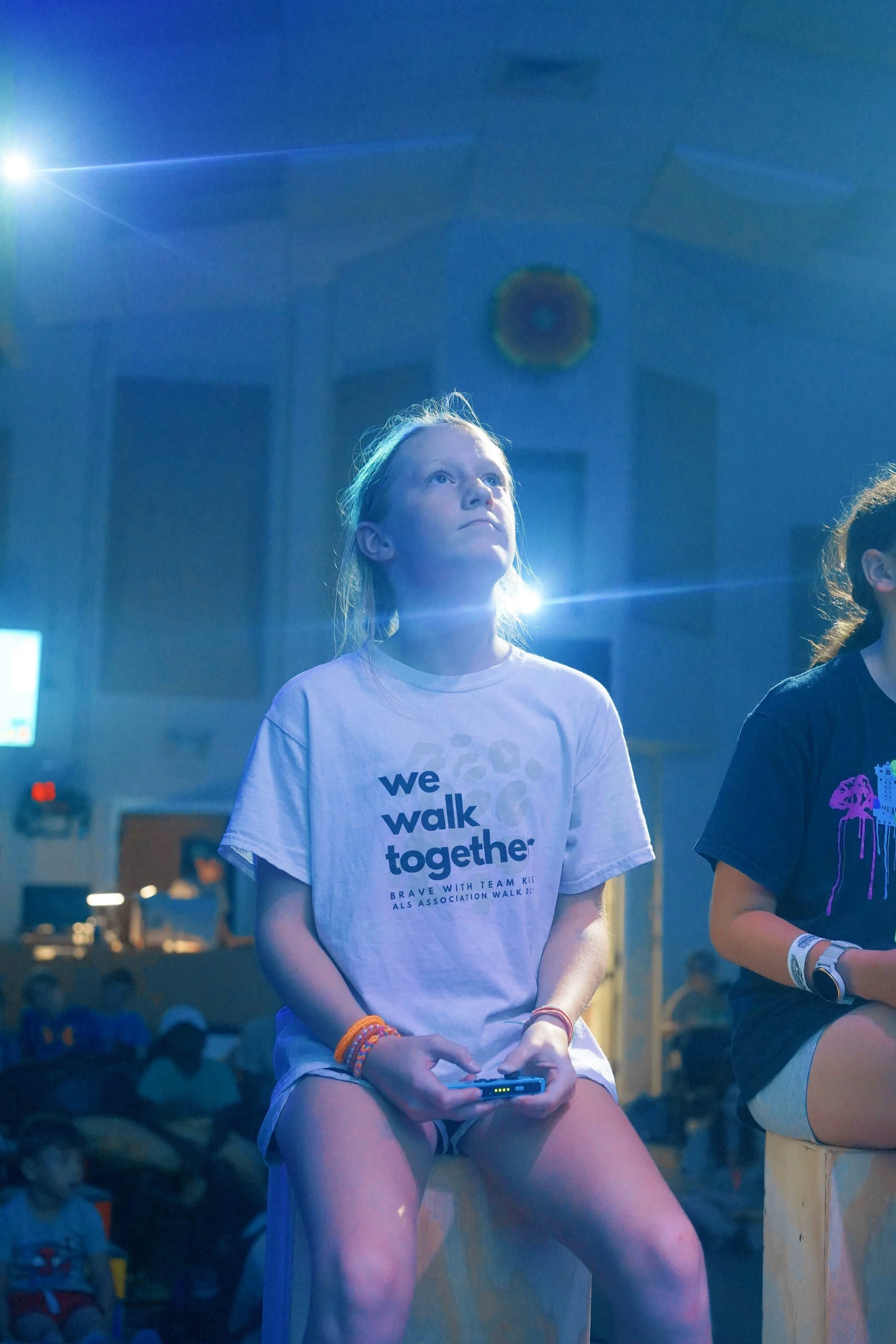 A young girl sitting on a wooden block at an indoor event, illuminated by blue stage lighting. She is wearing a white T-shirt that reads 'we walk together' and is holding a device in her hands. Other children are in the background, also sitting on wo