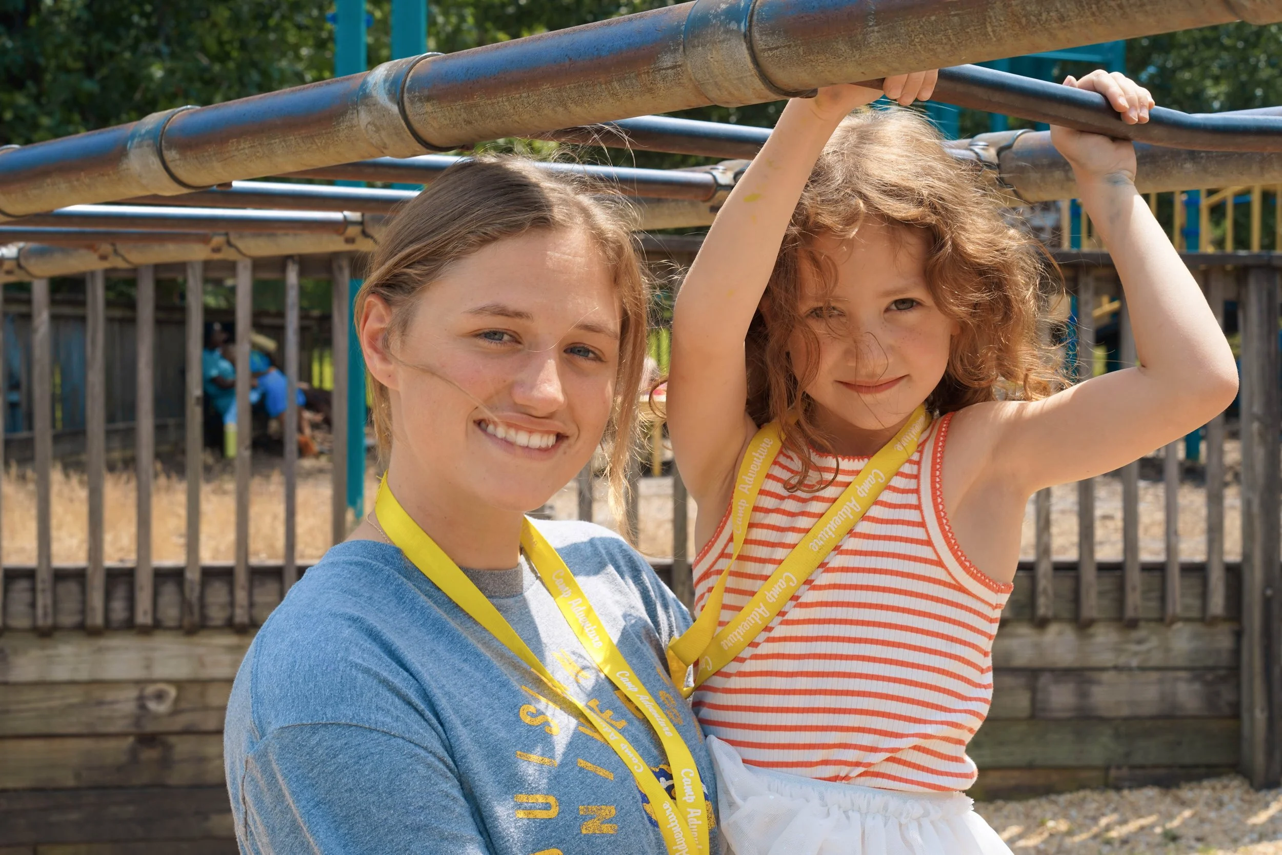 A young girl with curly red hair hanging from playground bars, smiling, with a woman smiling beside her at a playground.