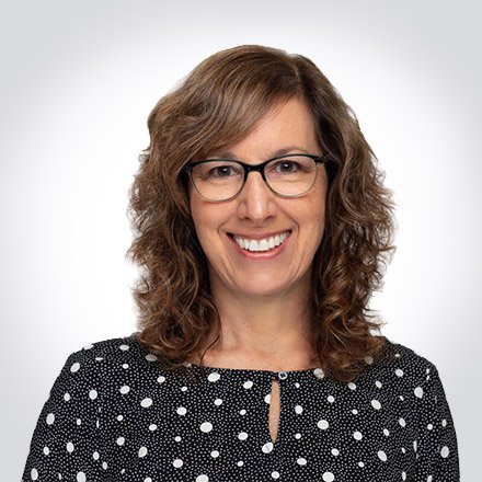 A woman with curly brown hair wearing glasses and a black polka dot blouse, smiling against a plain gray background.